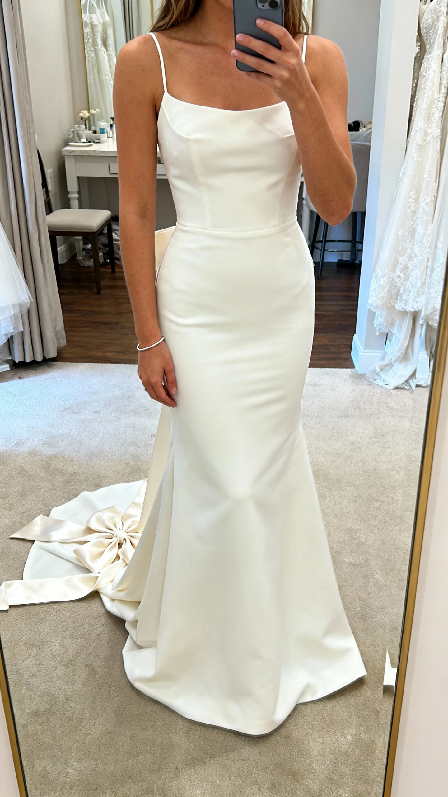 iPhone cropped-at-shoulders selfie showing a woman in a white crepe mermaid gown with a satin bow train cascading behind, simple bracelet, face not visible, bridal shop fitting room mirror, soft indoor lighting, natural iPhone photo.