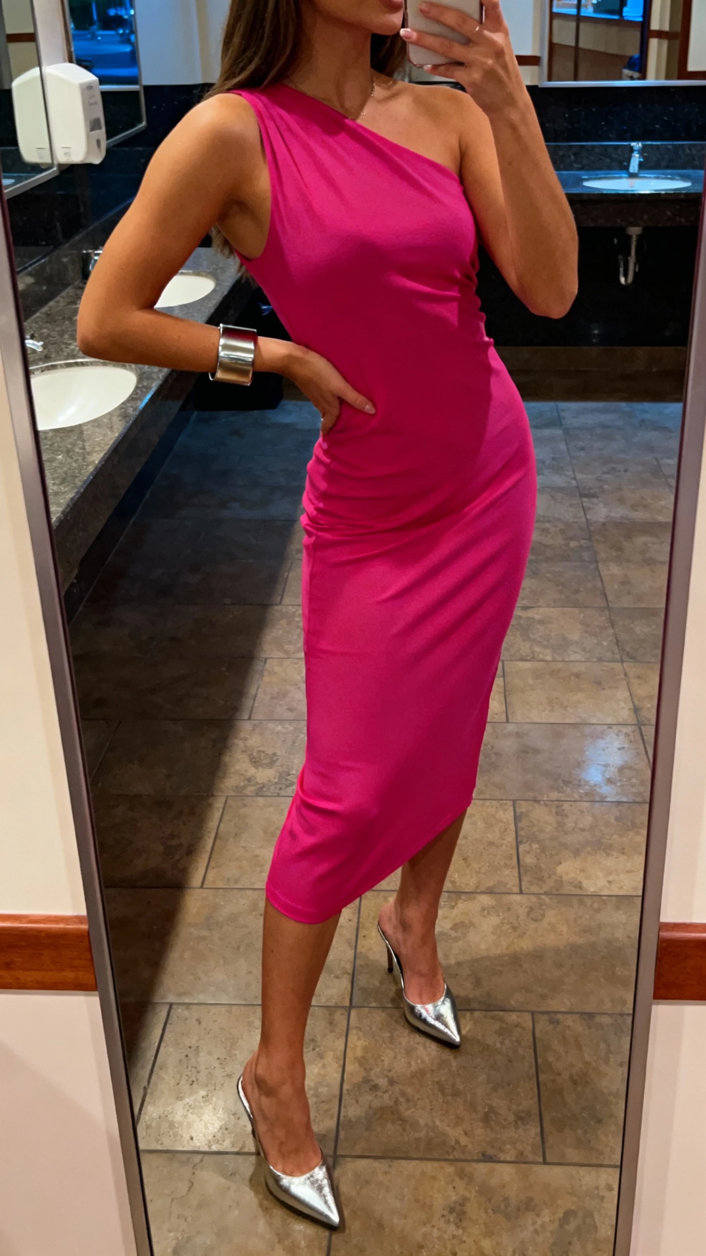 iPhone dance-floor mirror selfie of a woman in a bold-color one-shoulder midi dress, metallic heels, cuff bracelet, face not visible, side angle in venue restroom mirror, warm evening indoor light, natural iPhone photo.