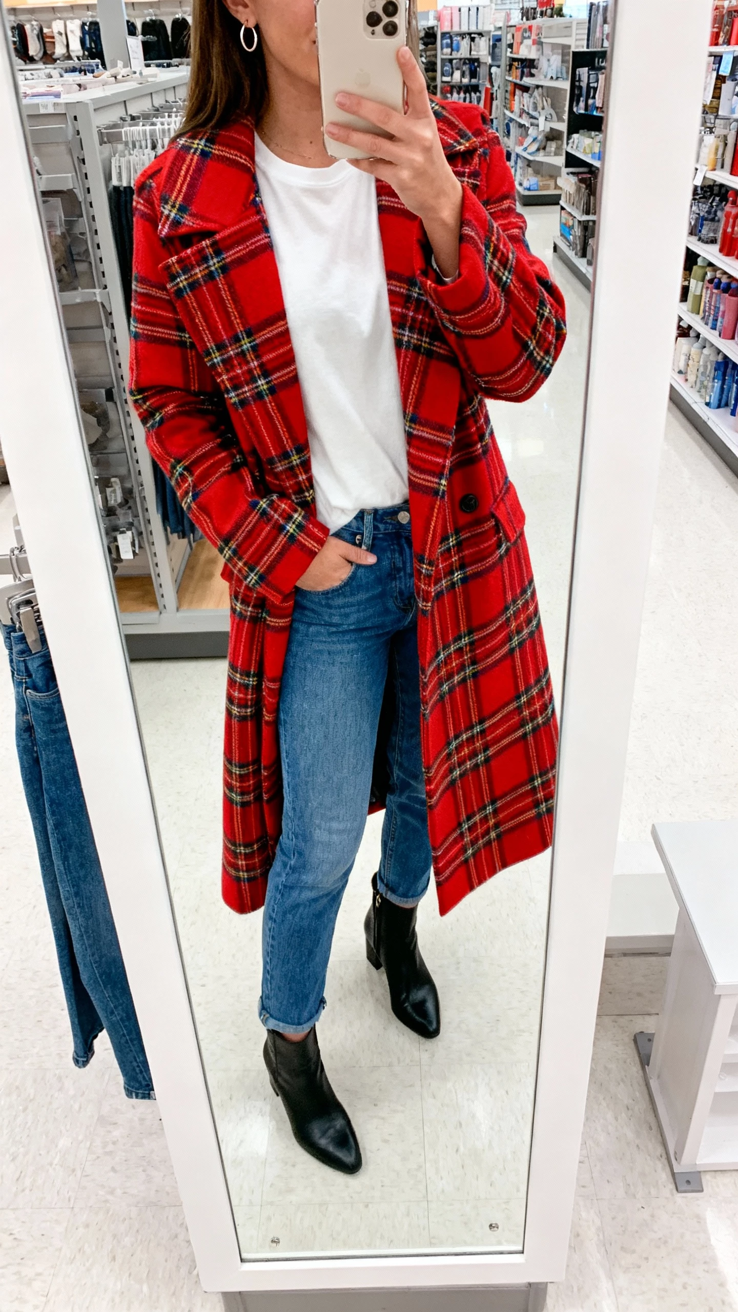 iPhone department-store mirror selfie of a woman wearing a bold statement coat (red or plaid) over simple basics: white tee, blue jeans, black ankle boots, small hoop earrings, face not visible, full-length mirror, bright indoor lighting, casual iPhone photo.