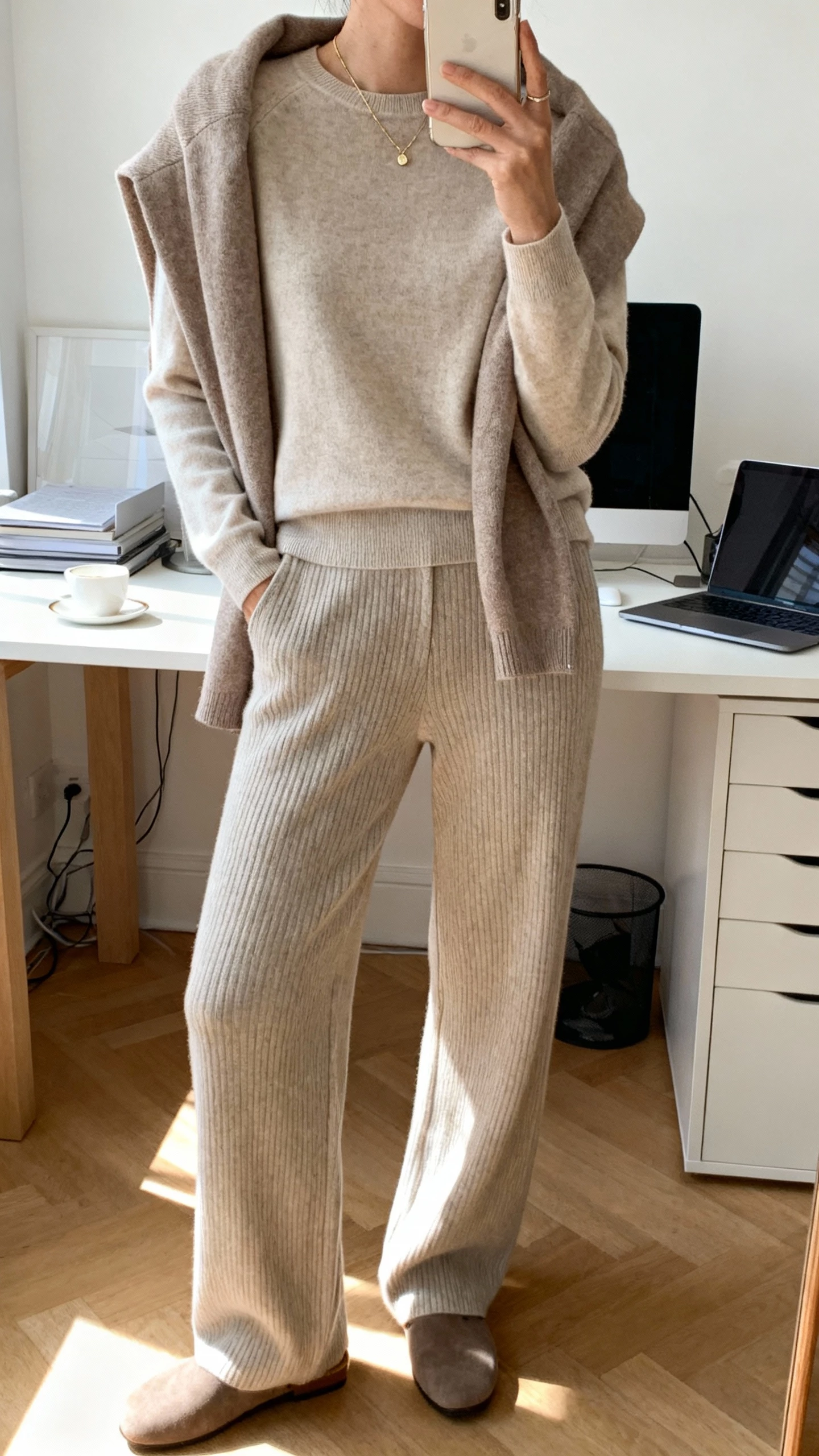 iPhone desk selfie of a woman in a work-from-anywhere cashmere capsule: cashmere crewneck, matching cardigan draped over shoulders, tailored knit pants, delicate necklace, and mule flats, face not visible, home office daylight, natural iPhone photo.