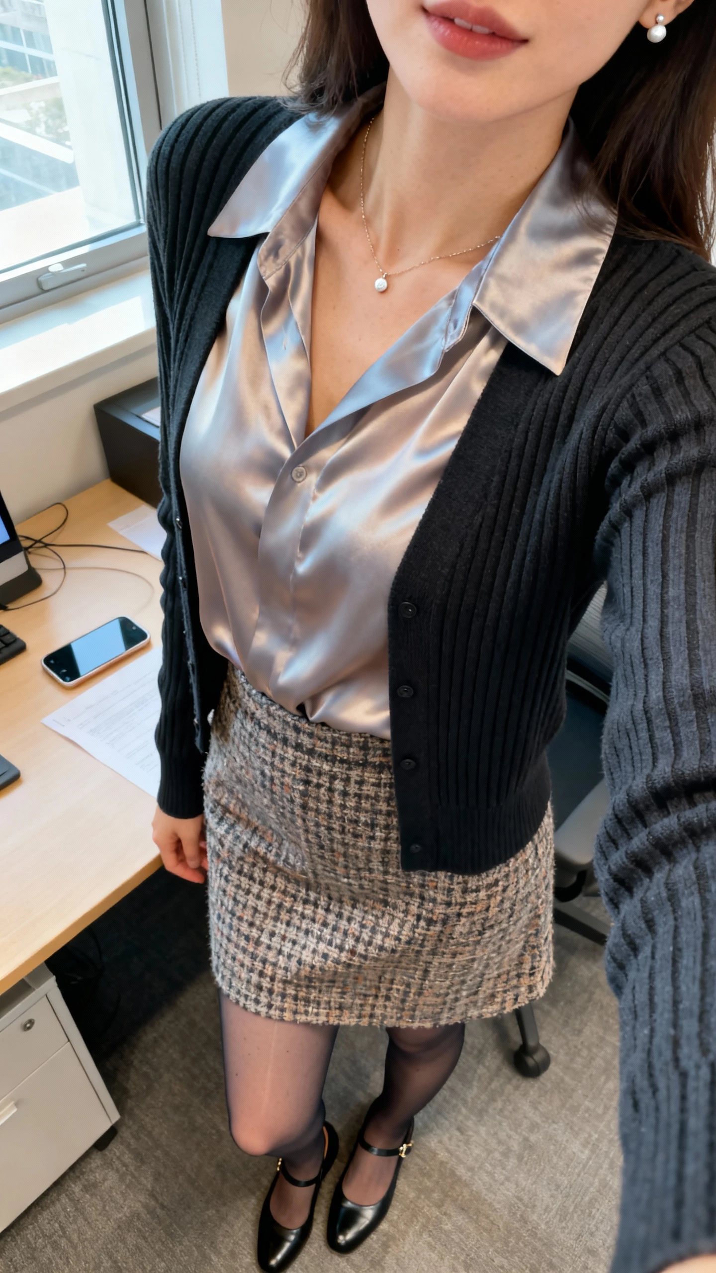 iPhone desk-side selfie cropped at shoulders showing a woman wearing a silk blouse under a ribbed cardigan with a tweed skirt, sheer tights, Mary Janes, delicate necklace and stud earrings, face not visible, office setting, window light, casual iPhone photo.