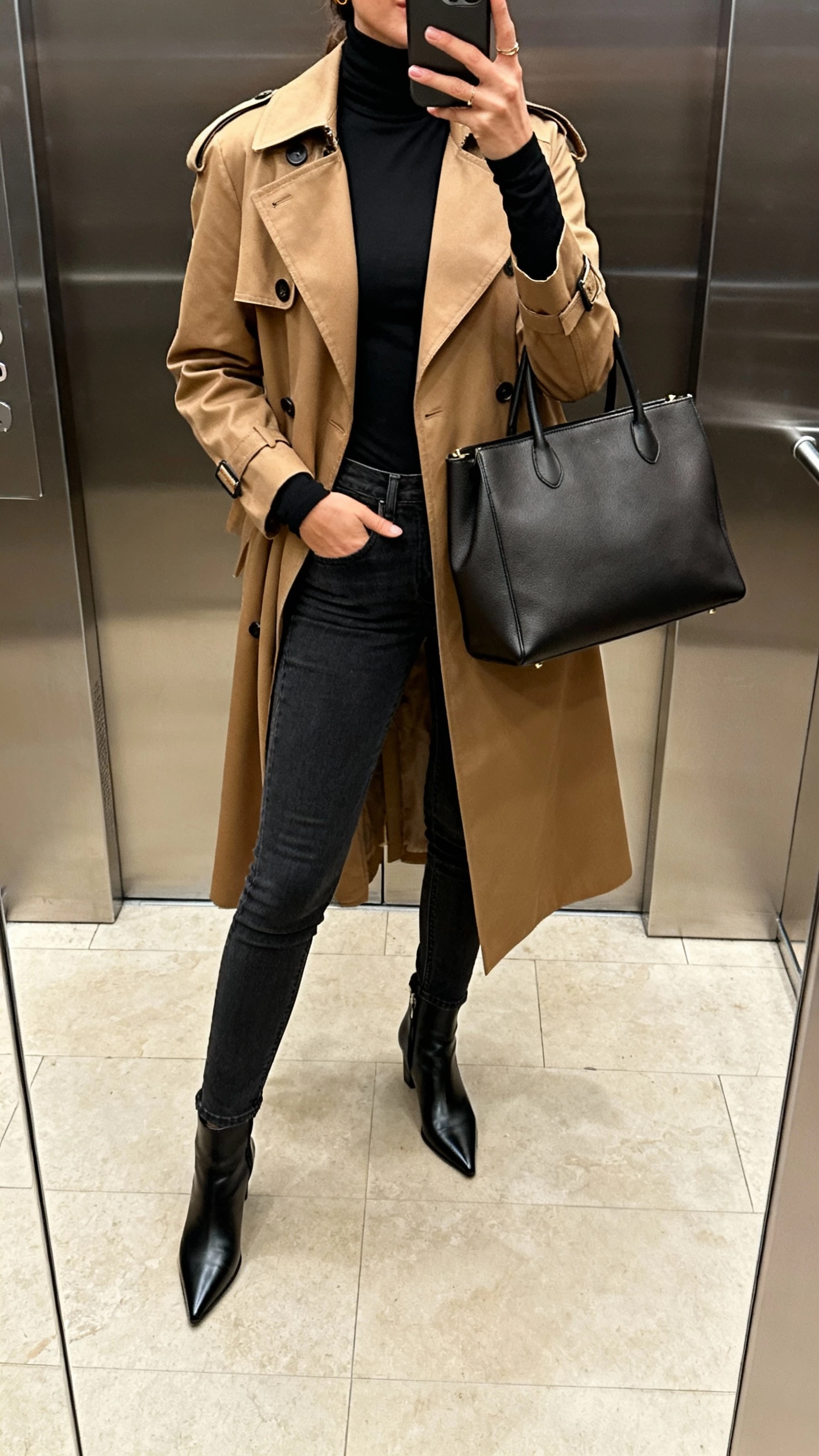 iPhone elevator mirror selfie of a woman in a black fitted turtleneck, camel trench coat, slim dark jeans, pointed ankle boots, structured tote, face not visible, indoor lighting, natural iPhone photo quality.