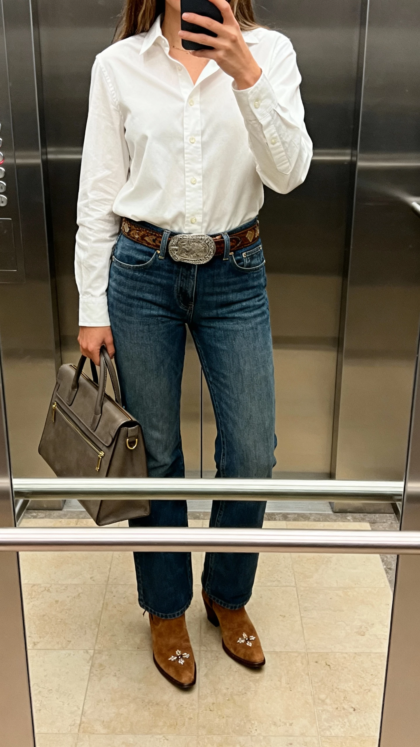 iPhone elevator mirror selfie of a woman in a crisp button-down shirt tucked into bootcut jeans, Western-style belt, suede ankle boots, subtle concho detail and a structured work tote; face not visible, cropped at shoulders, indoor lighting, natural iPhone photo quality.