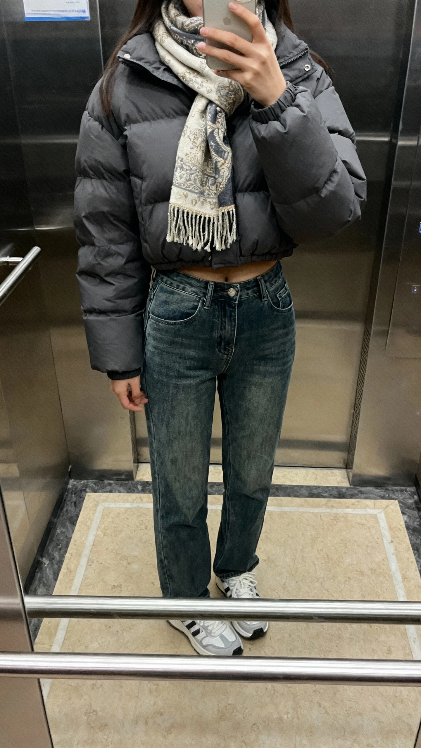 iPhone elevator mirror selfie of a woman in a cropped puffer jacket, straight-leg jeans, an elegant scarf, and clean sneakers, face not visible, indoor elevator lighting, natural iPhone photo.