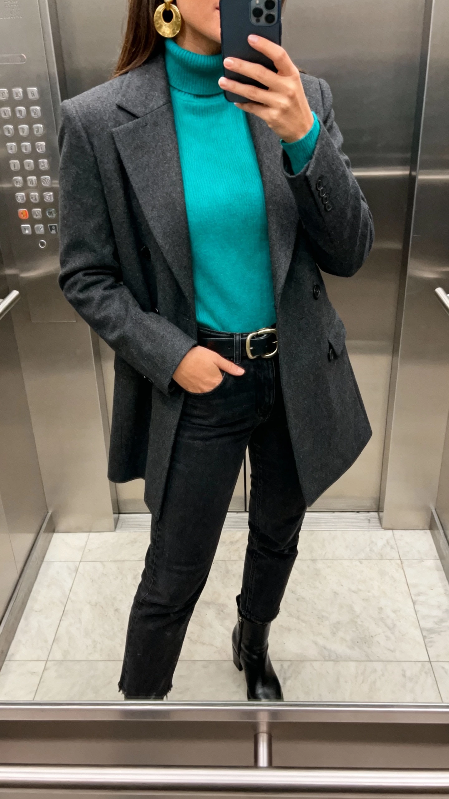 iPhone elevator-mirror selfie of a woman in a teal turtleneck, charcoal tailored coat, black jeans, burnished brass statement earrings and buckle belt, black ankle boots, face not visible, indoor lighting, natural iPhone photo