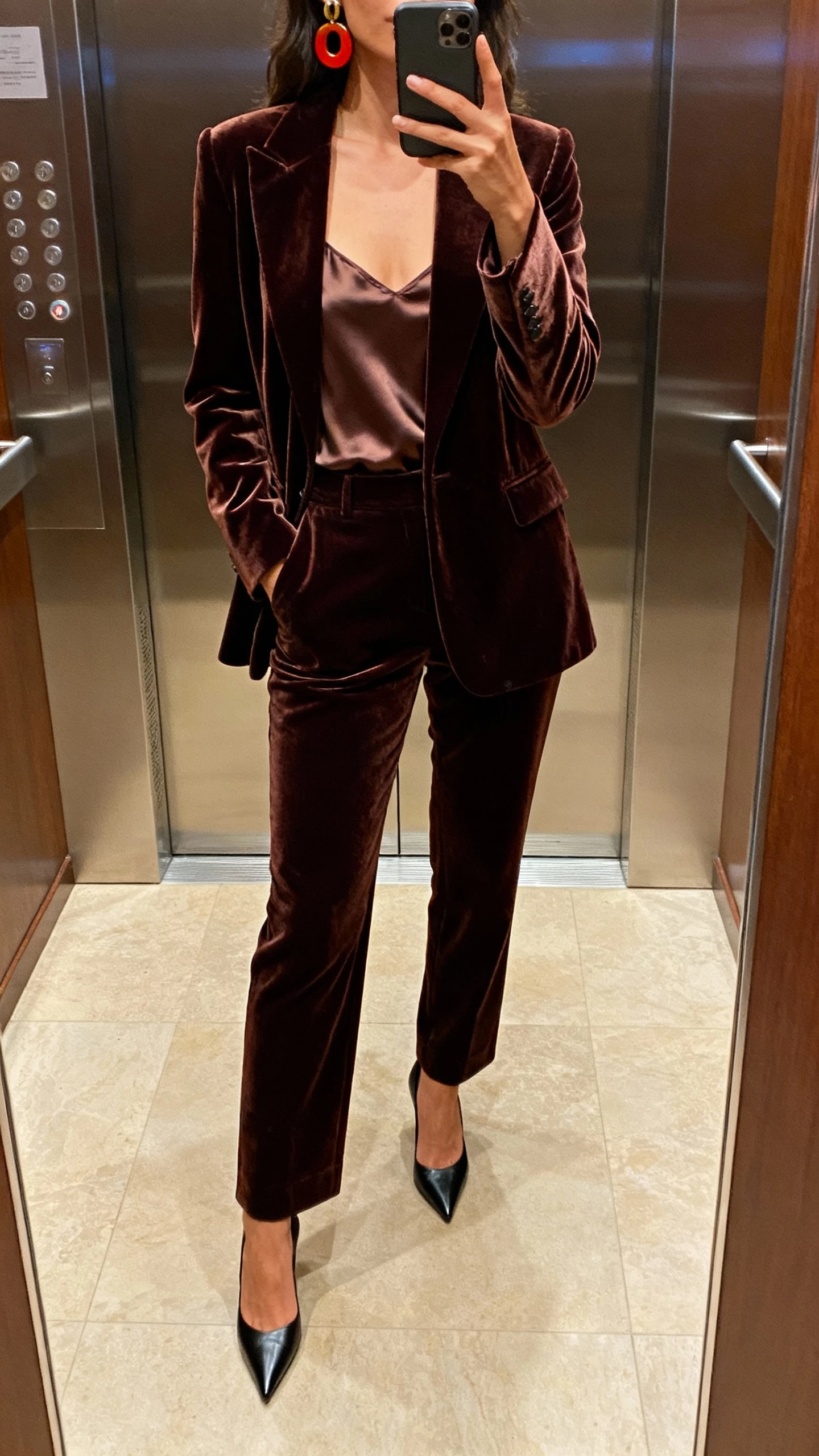 iPhone elevator mirror selfie of a woman in a velvet blazer and matching tailored trousers set, silky camisole underneath, pointed heels and bold earrings, face not visible, evening hallway lighting, natural iPhone photo quality.