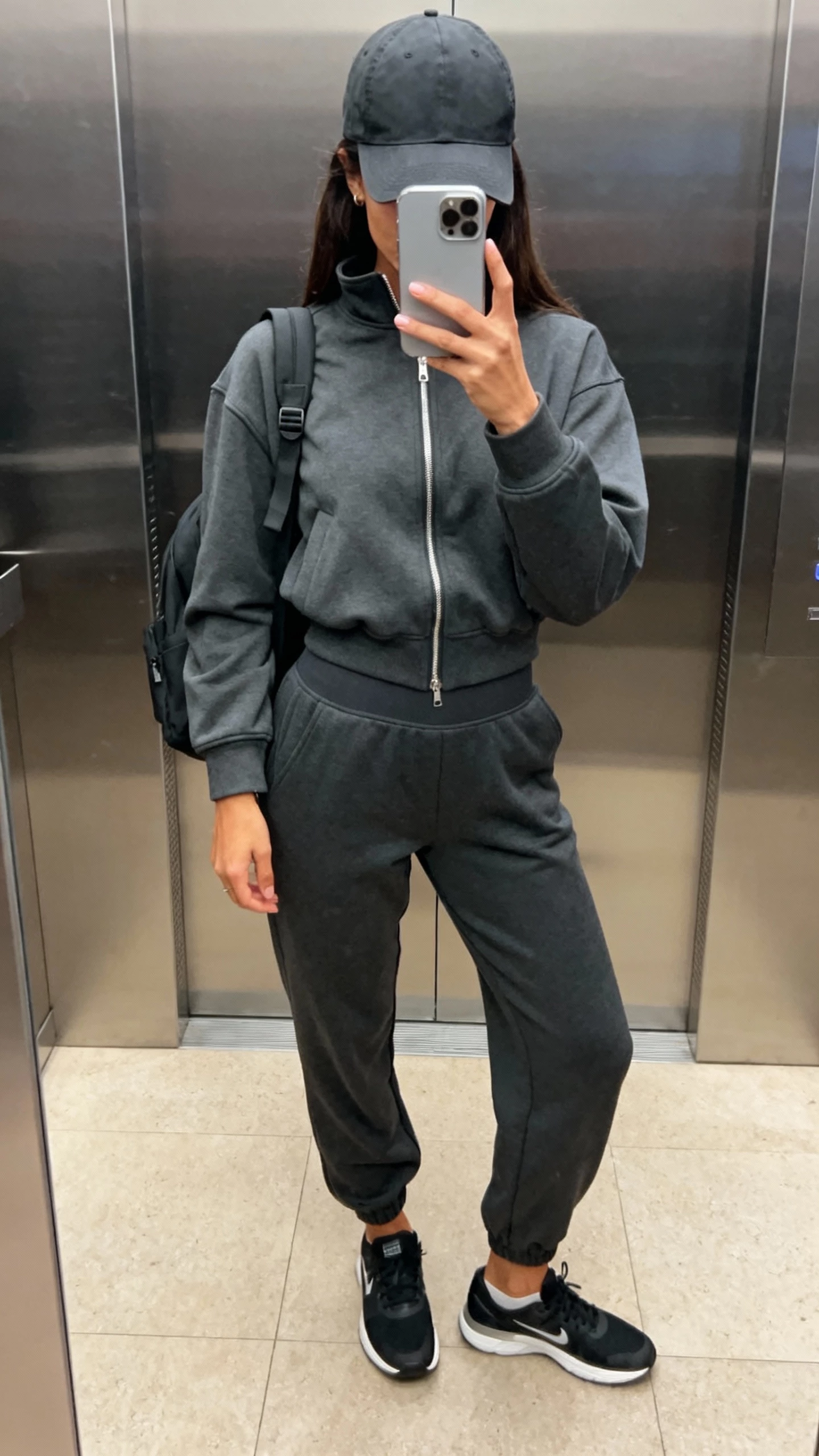 iPhone elevator mirror selfie of a woman in monochrome athleisure: charcoal zip-up track jacket, matching high-waist joggers, sleek trainers, structured cap, minimalist backpack, face not visible, indoor lighting, iPhone photo quality.