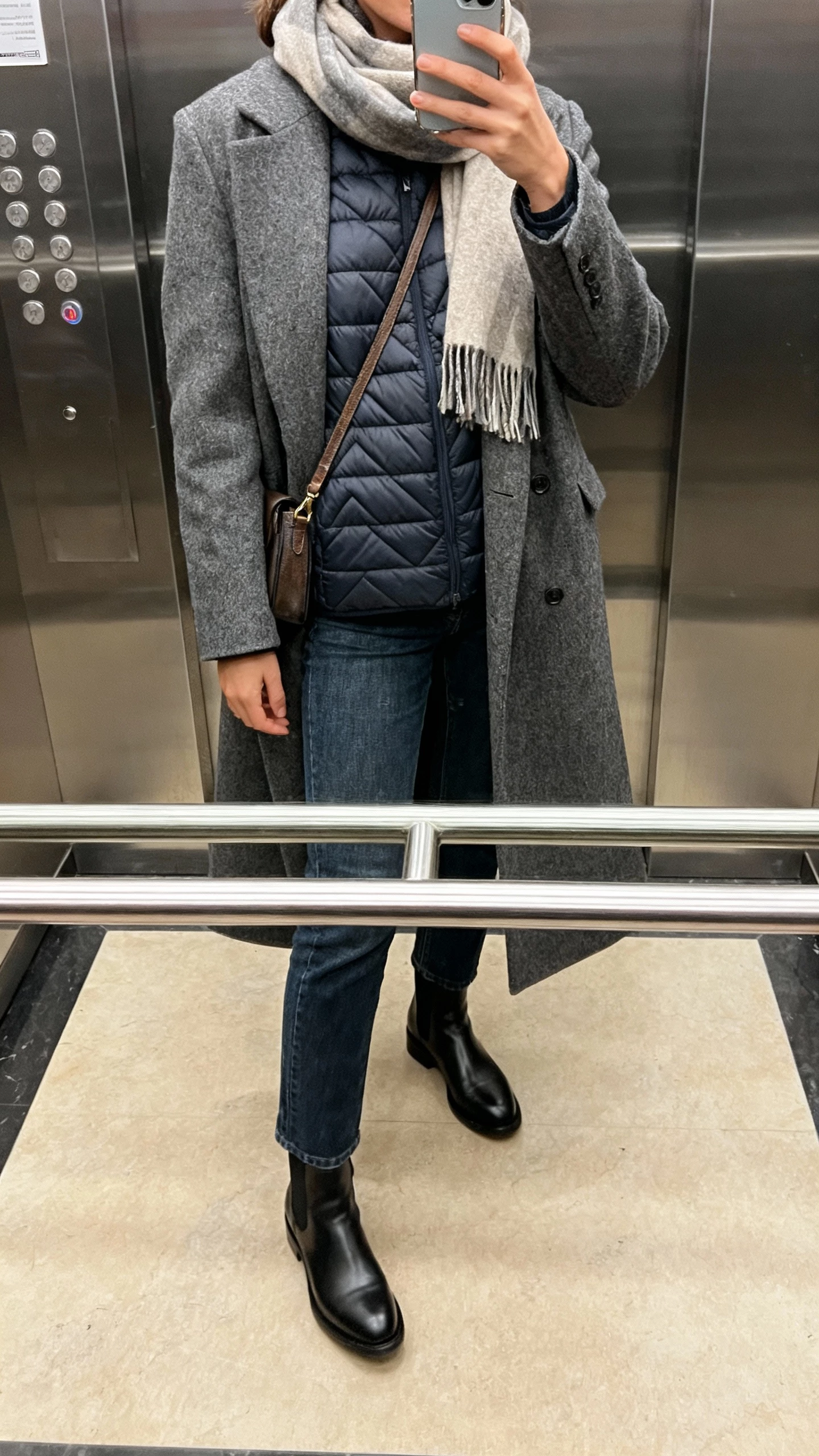 iPhone elevator-mirror selfie of a woman wearing a thin quilted puffer under a long wool coat, straight jeans, Chelsea boots, scarf and crossbody, face not visible, cropped at neck, natural indoor lighting, natural iPhone photo quality.