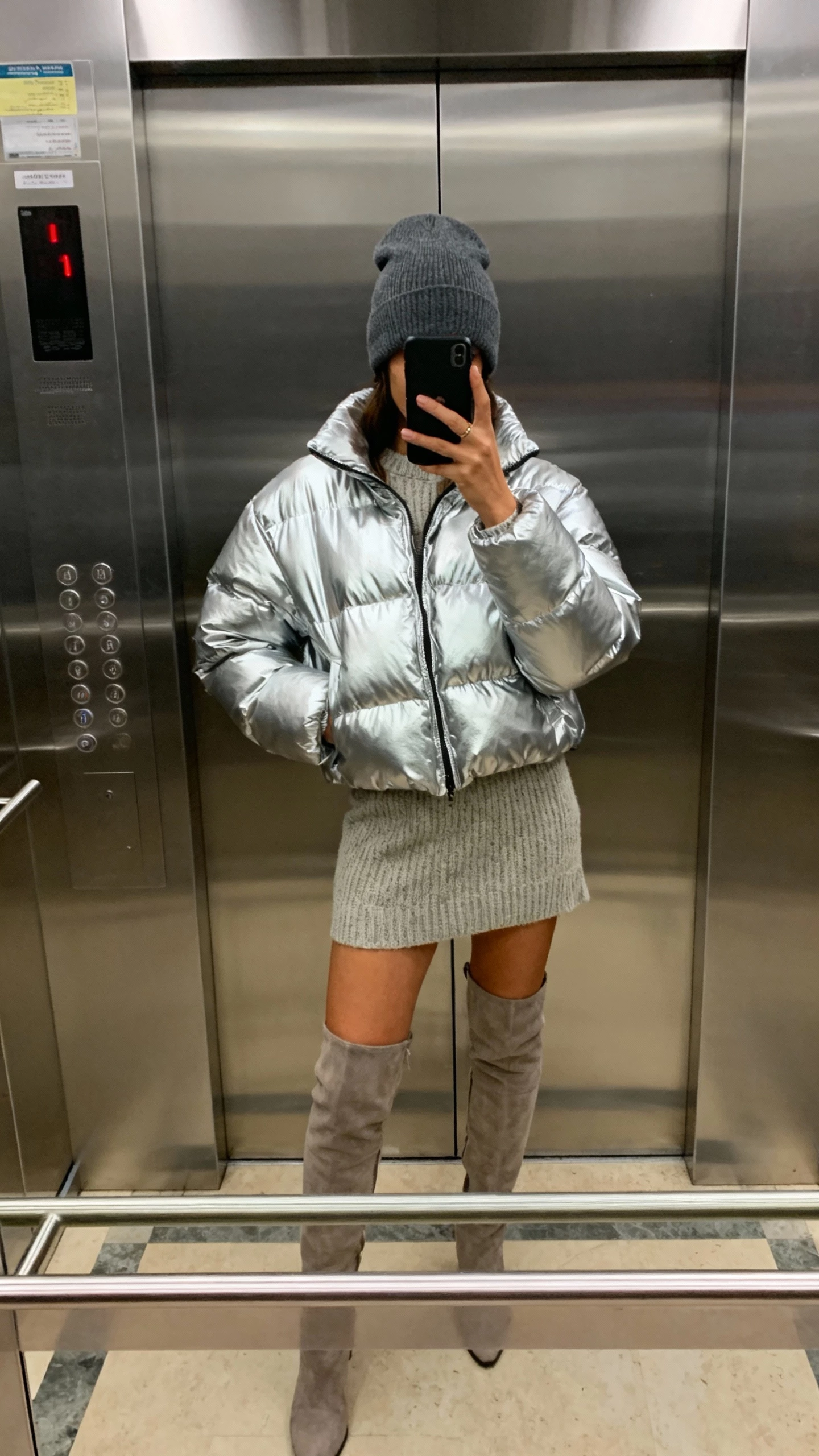 iPhone elevator mirror selfie of a woman wearing a metallic puffer, knit mini skirt, over-the-knee boots, and a beanie, face not visible, mixed indoor lighting, casual iPhone photo quality.