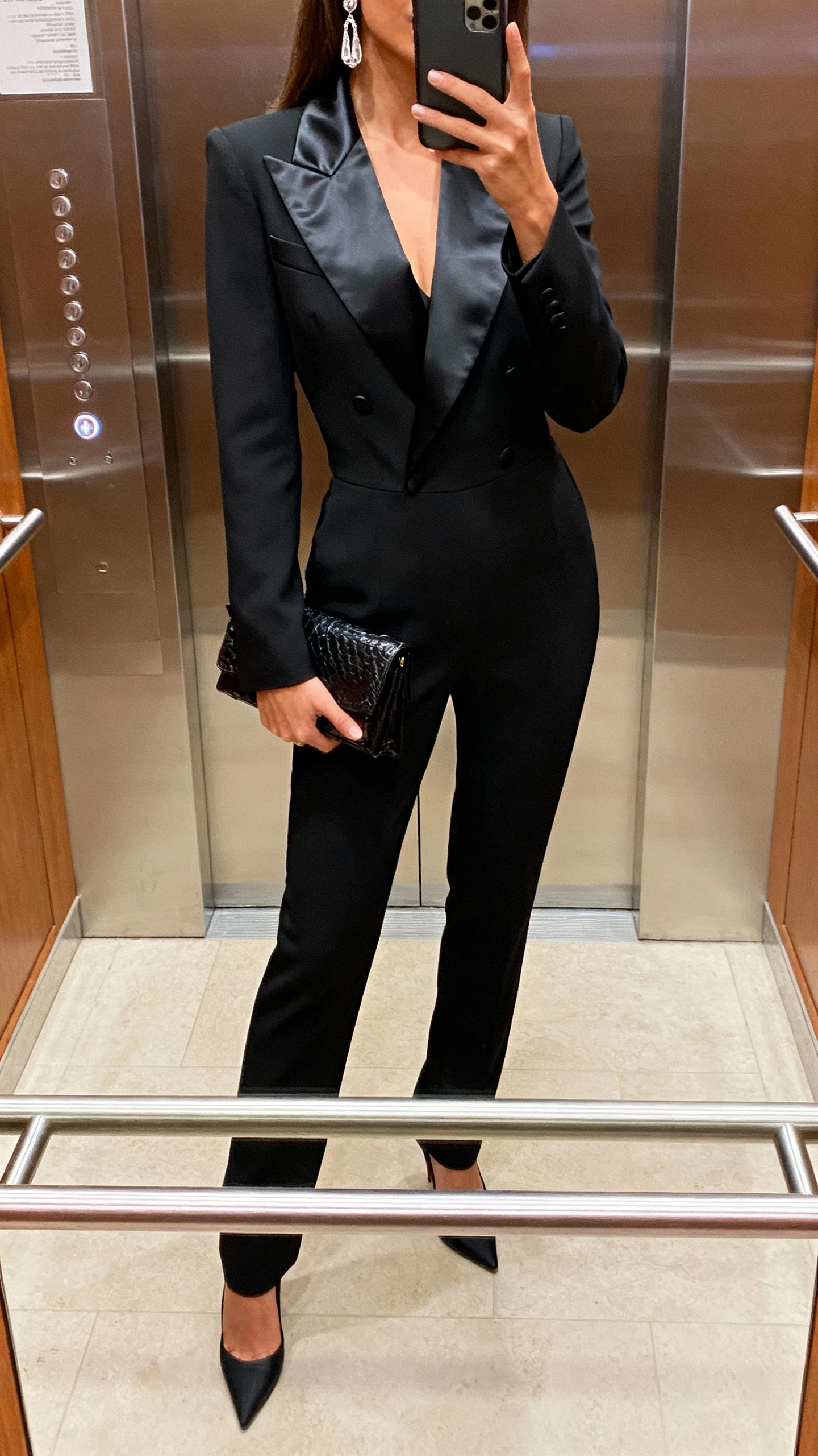 iPhone elevator mirror selfie of a woman wearing a tuxedo-inspired jumpsuit with satin lapels, sleek heels, clutch, drop earrings, face not visible, cropped at shoulders, soft indoor lighting, natural iPhone photo quality