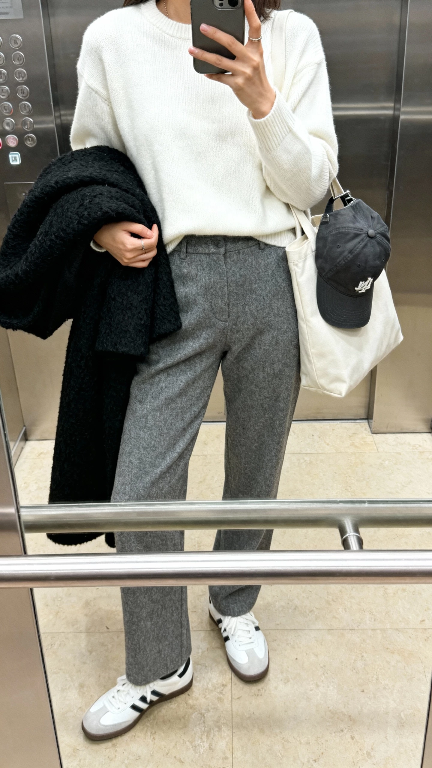 iPhone elevator mirror selfie of a woman wearing grey wool trousers, white crewneck knit, classic white sneakers, black wool coat over arm, baseball cap clipped to tote, face not visible, indoor lighting, natural iPhone photo quality.