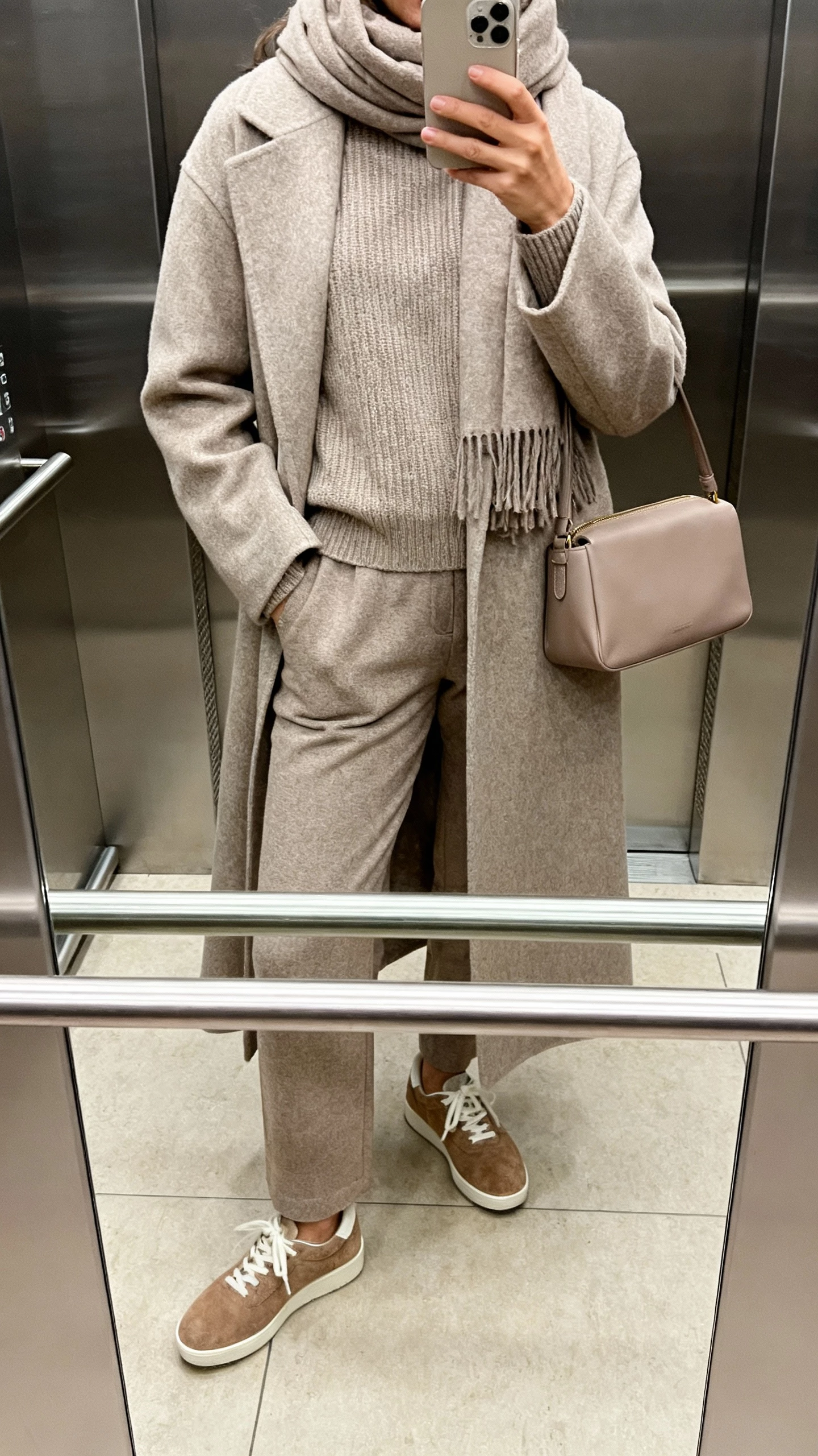 iPhone elevator mirror selfie of a woman wearing layered monochrome pieces: long wool coat over a tonal knit, matching trousers, and suede sneakers, with a scarf and structured bag in the same shade; face not visible, soft indoor lighting, natural iPhone photo quality.