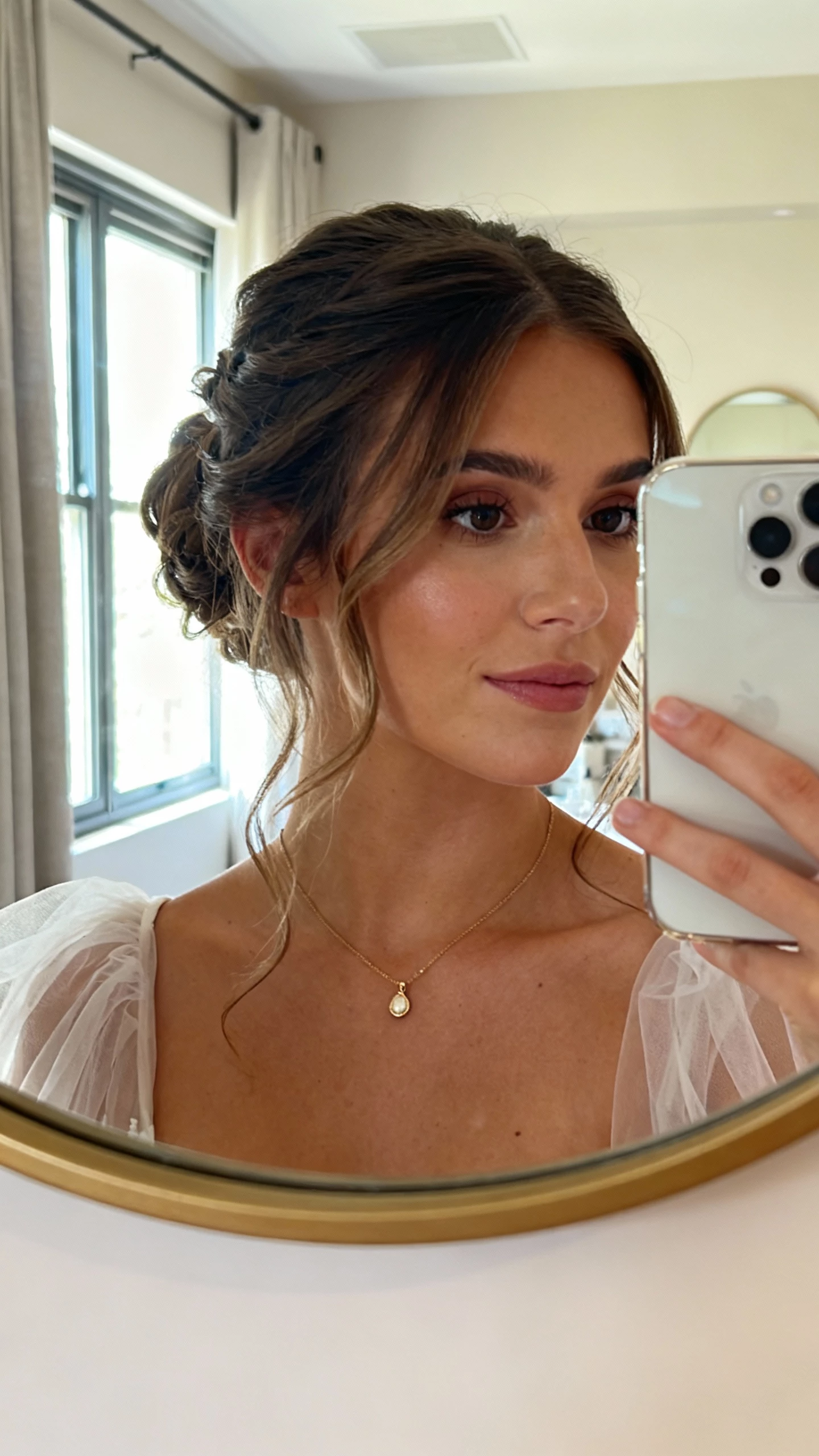 iPhone front mirror selfie cropped at nose level showing a half-up illusion updo with loose romantic pieces, simple pendant necklace, tulle sleeves; face not visible, bridal suite mirror, window light, natural iPhone aesthetic.