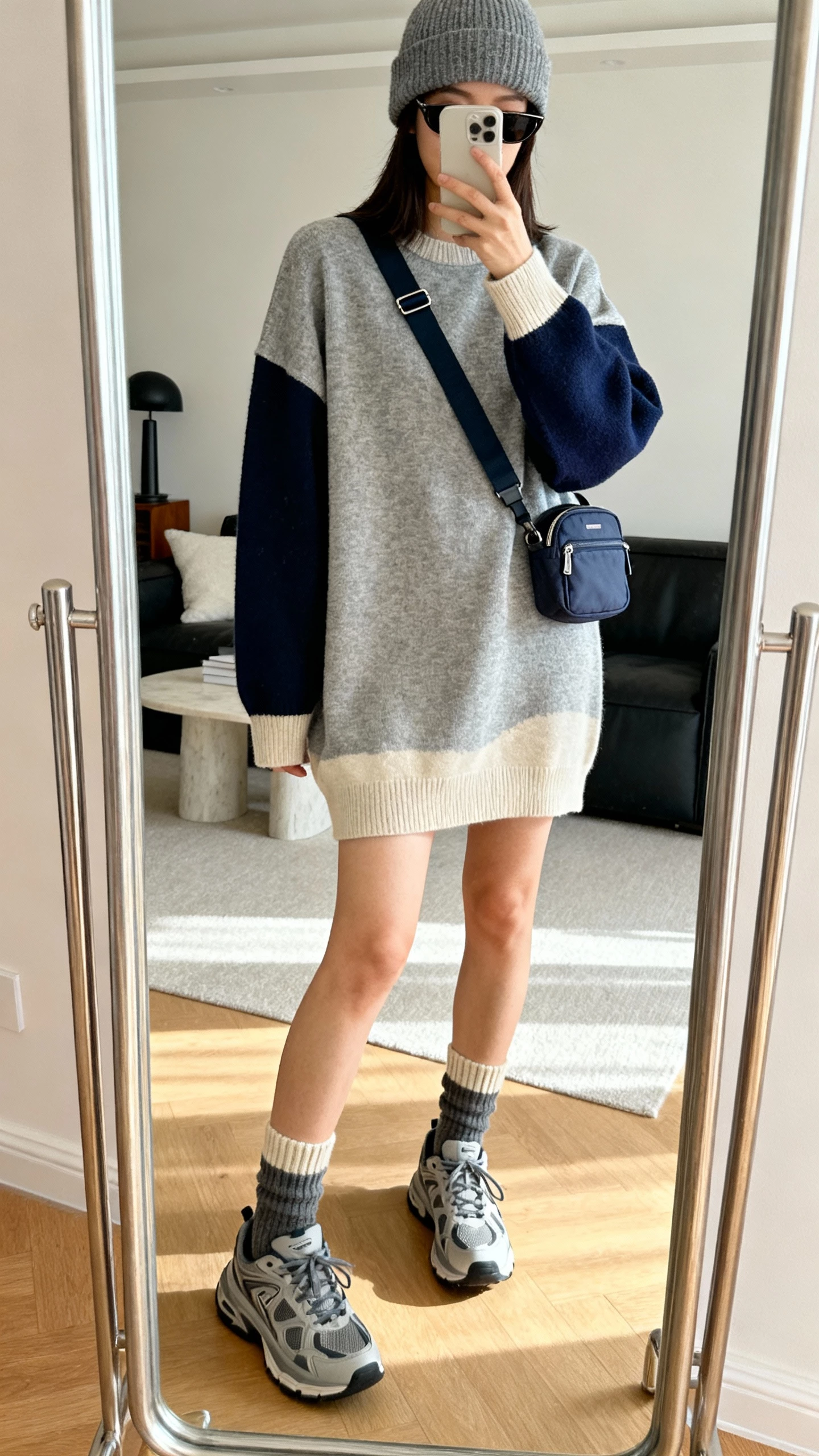 iPhone front mirror selfie of a woman in a boxy crewneck sweater dress (heather gray/oatmeal/navy) with chunky sneakers, ribbed ankle socks peeking, casual crossbody belt bag or mini backpack, cozy beanie, slim sunglasses; face not visible, cropped at shoulders, living room full-length mirror, soft daylight, natural iPhone photo quality.