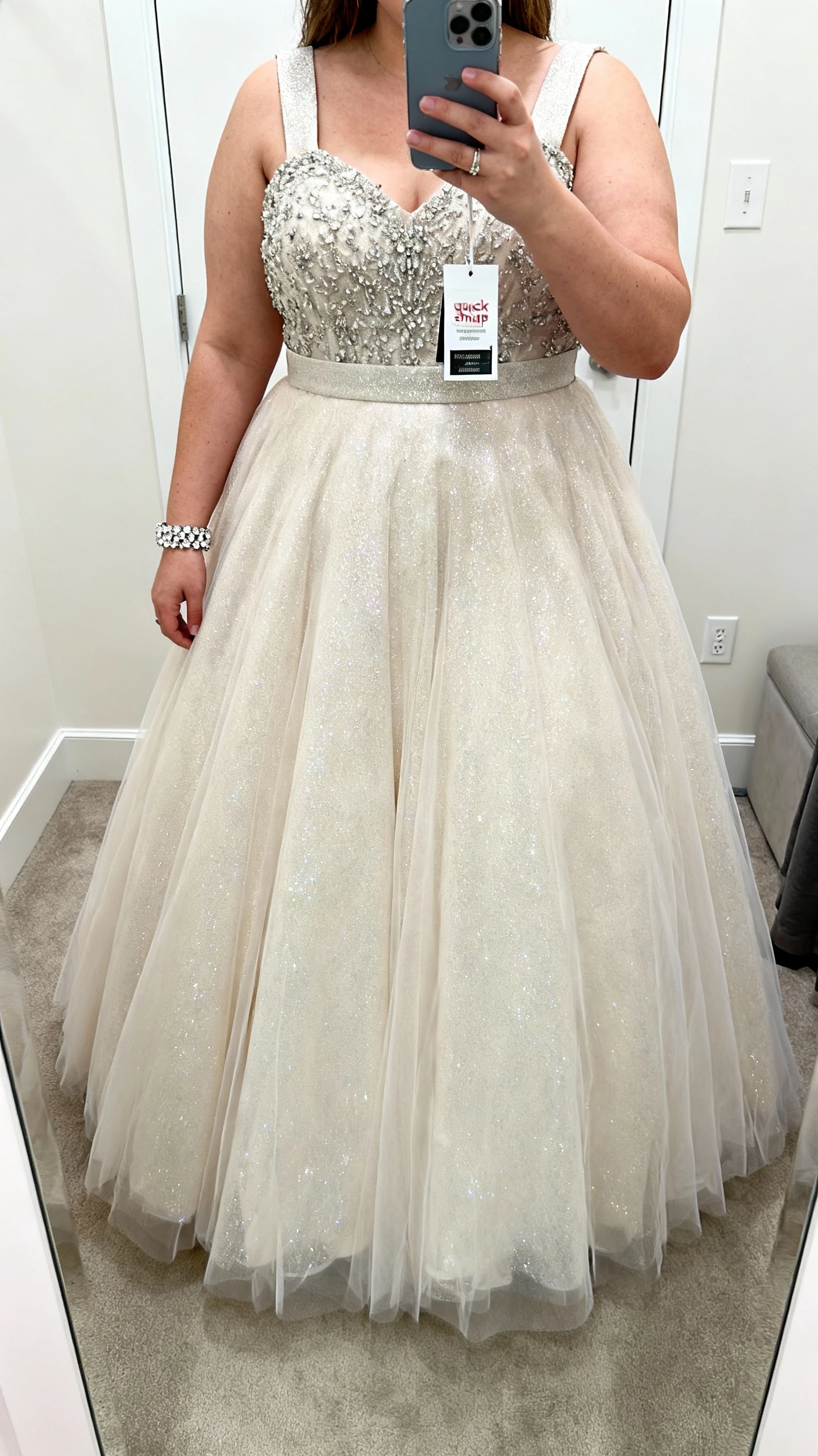 iPhone full-length mirror selfie of a woman in David’s Bridal Plus (Oleg Cassini & Galina Signature) gown: glitter tulle ballgown with beaded bodice and wide straps, quick-ship tag still on hanger, styled with a crystal bracelet; face not visible, fitting room mirror, bright indoor lighting, natural iPhone photo quality.