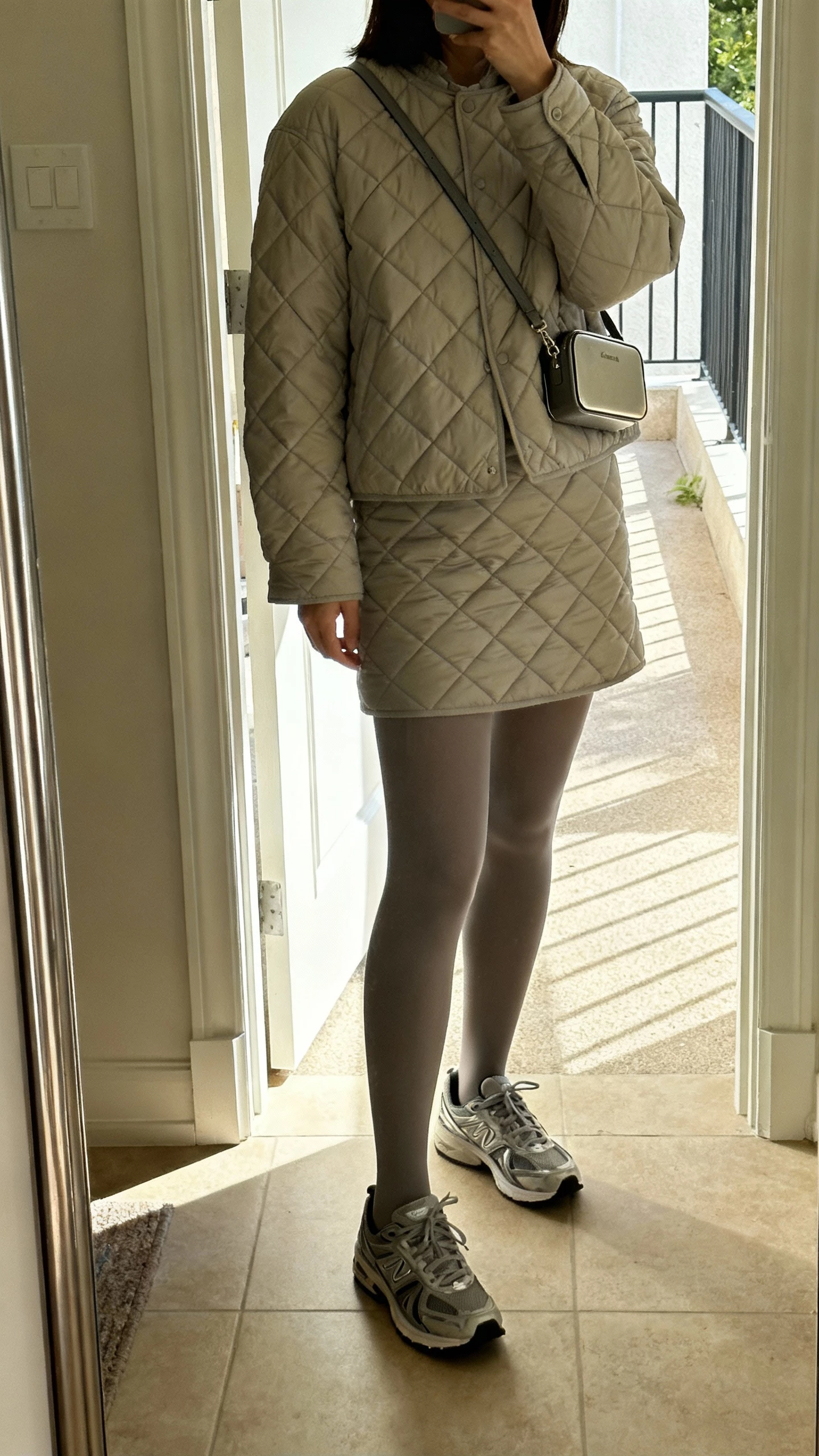 iPhone full-length mirror selfie of a woman wearing a quilted skirt set (quilted jacket and matching quilted skirt) with opaque tights, sleek sneakers, and a compact crossbody; face not visible, entryway mirror with daylight, natural iPhone photo.