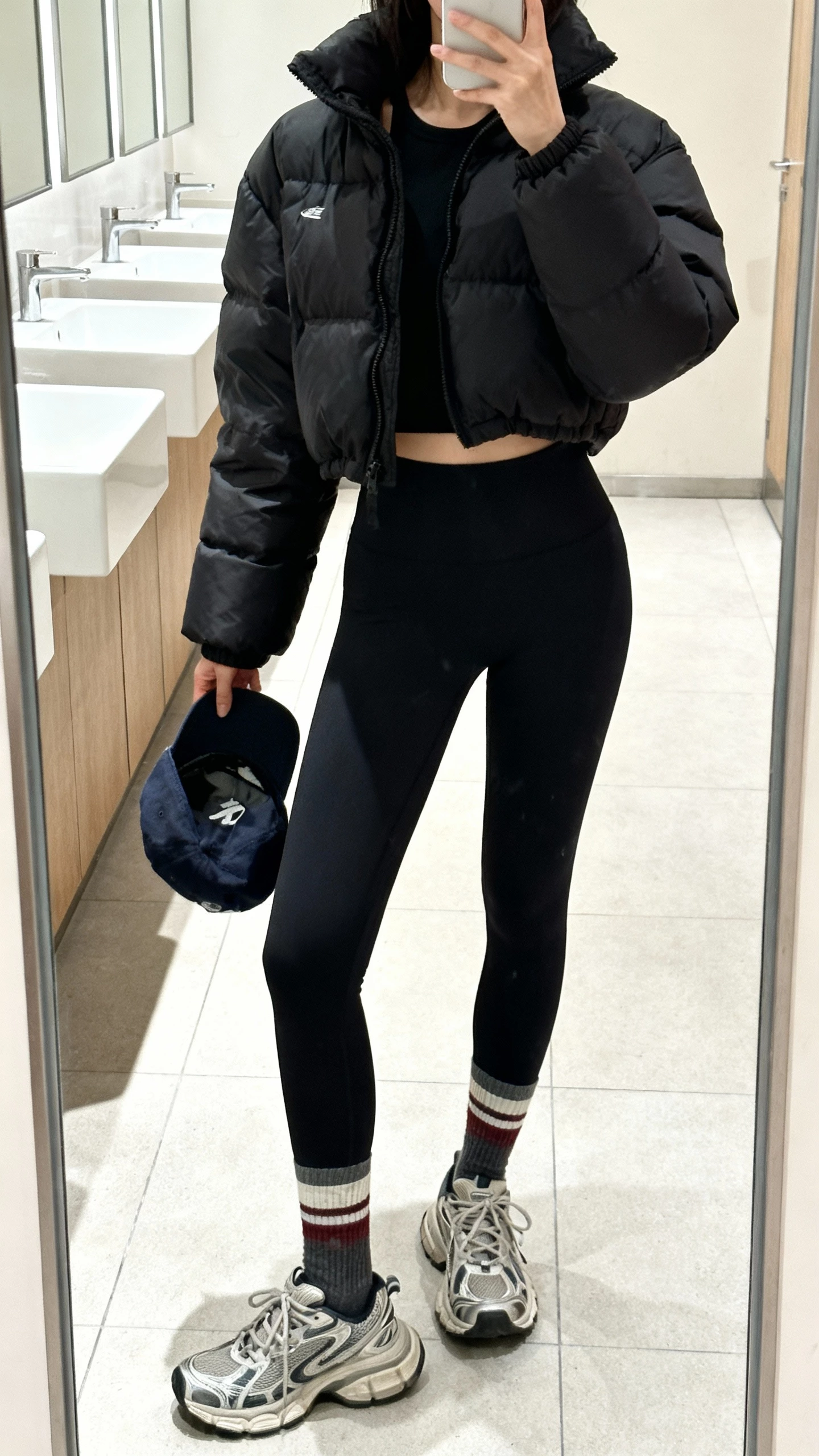 iPhone gym-bathroom mirror selfie of a woman wearing a black puffer jacket, high-rise black leggings, sporty crew socks, chunky sneakers, baseball cap in hand, face not visible, full-length mirror, bright indoor lighting, casual iPhone photo.