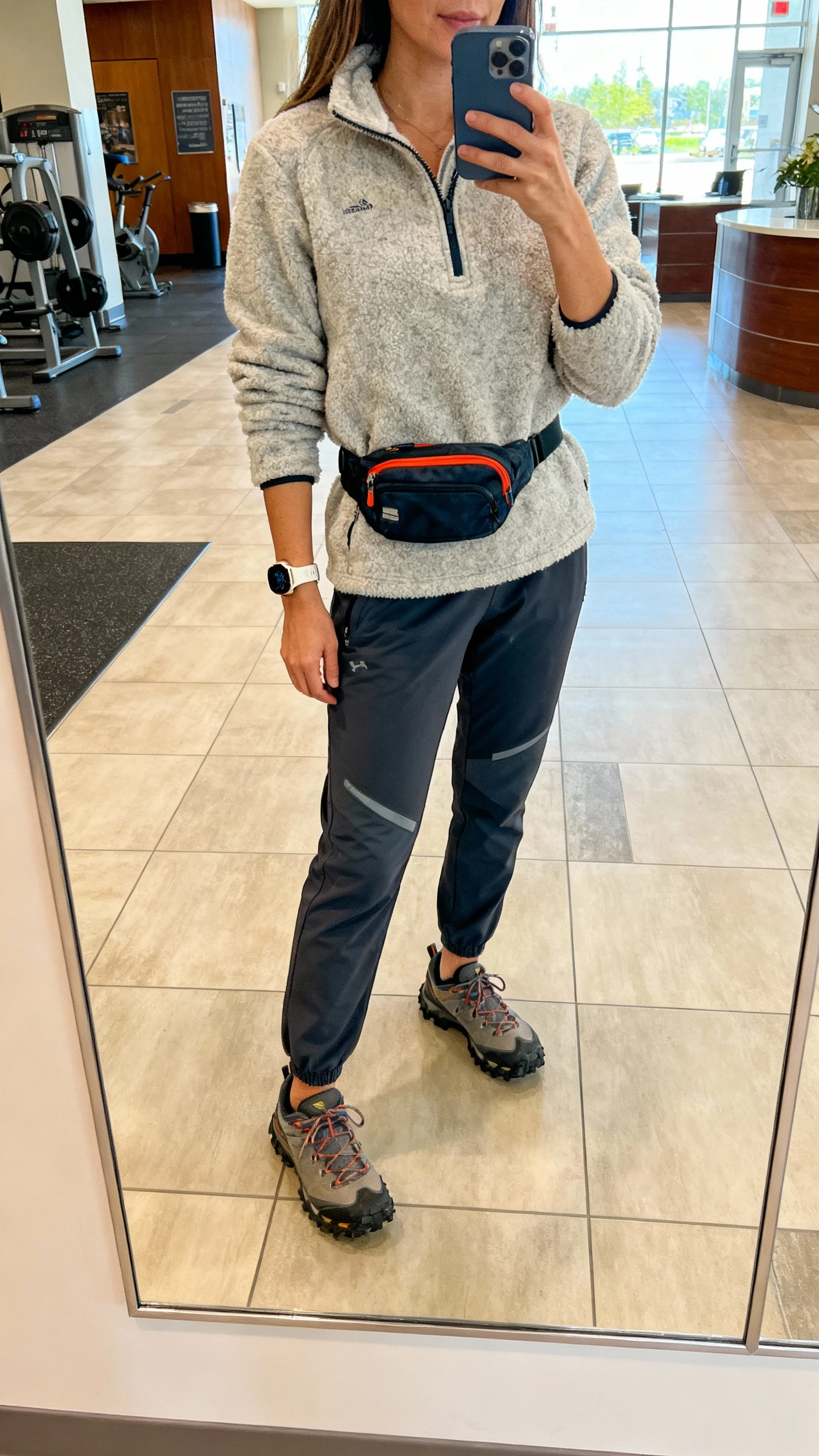 iPhone gym-lobby mirror selfie of a woman in a fleece half-zip, tech pants, trail sneakers, sporty watch, belt bag, face not visible, bright indoor lighting, natural iPhone photo quality.