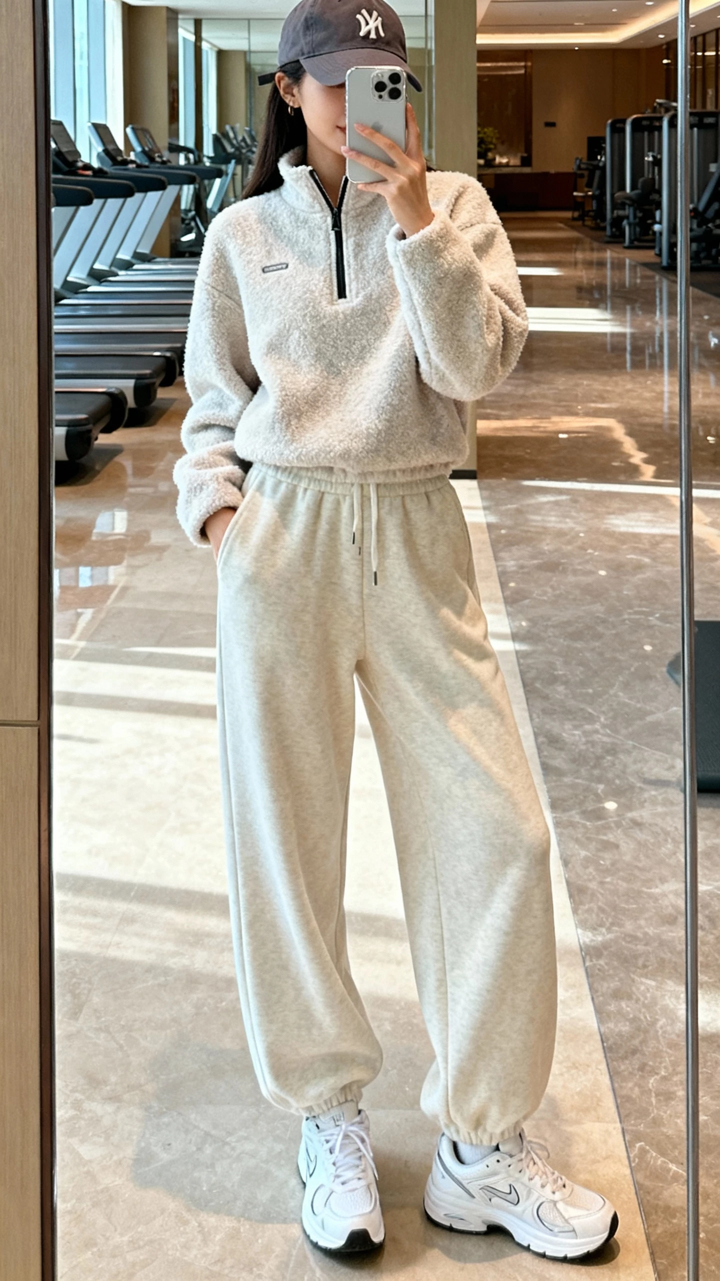 iPhone gym-lobby mirror selfie of a woman in a fleece half-zip, wide-leg joggers, sleek white sneakers, and a baseball cap, face not visible, cropped at shoulders, bright indoor light, casual iPhone photo.