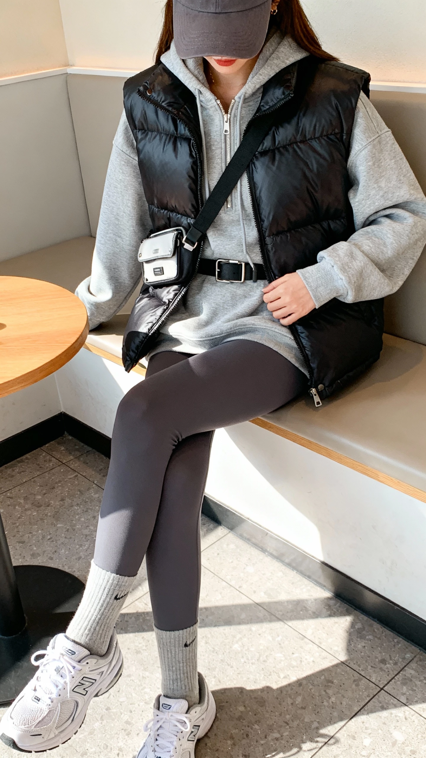 iPhone gym-to-brunch selfie of a woman wearing a sleek black puffer vest layered over a heather gray zip hoodie and matching charcoal leggings, baseball cap, crew socks with white sneakers, belt bag worn crossbody, face not visible, taken in a bright cafe corner seat, natural daylight, casual iPhone photo