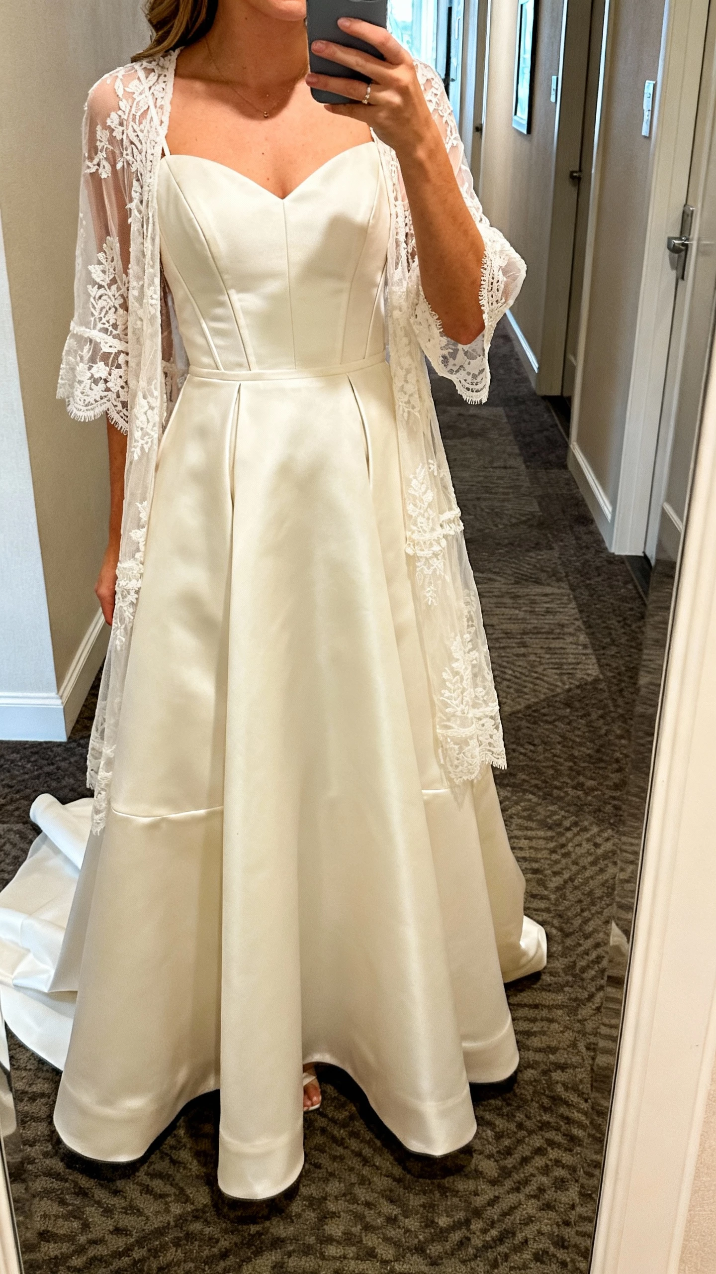 iPhone half-body selfie of a woman wearing a Justin Alexander Signature clean satin fit-and-flare with sculpted seams and detachable overskirt, layered with a Lillian West sheer lace topper for boho vibe; face not visible, hotel hallway mirror, soft indoor lighting, natural iPhone shot.