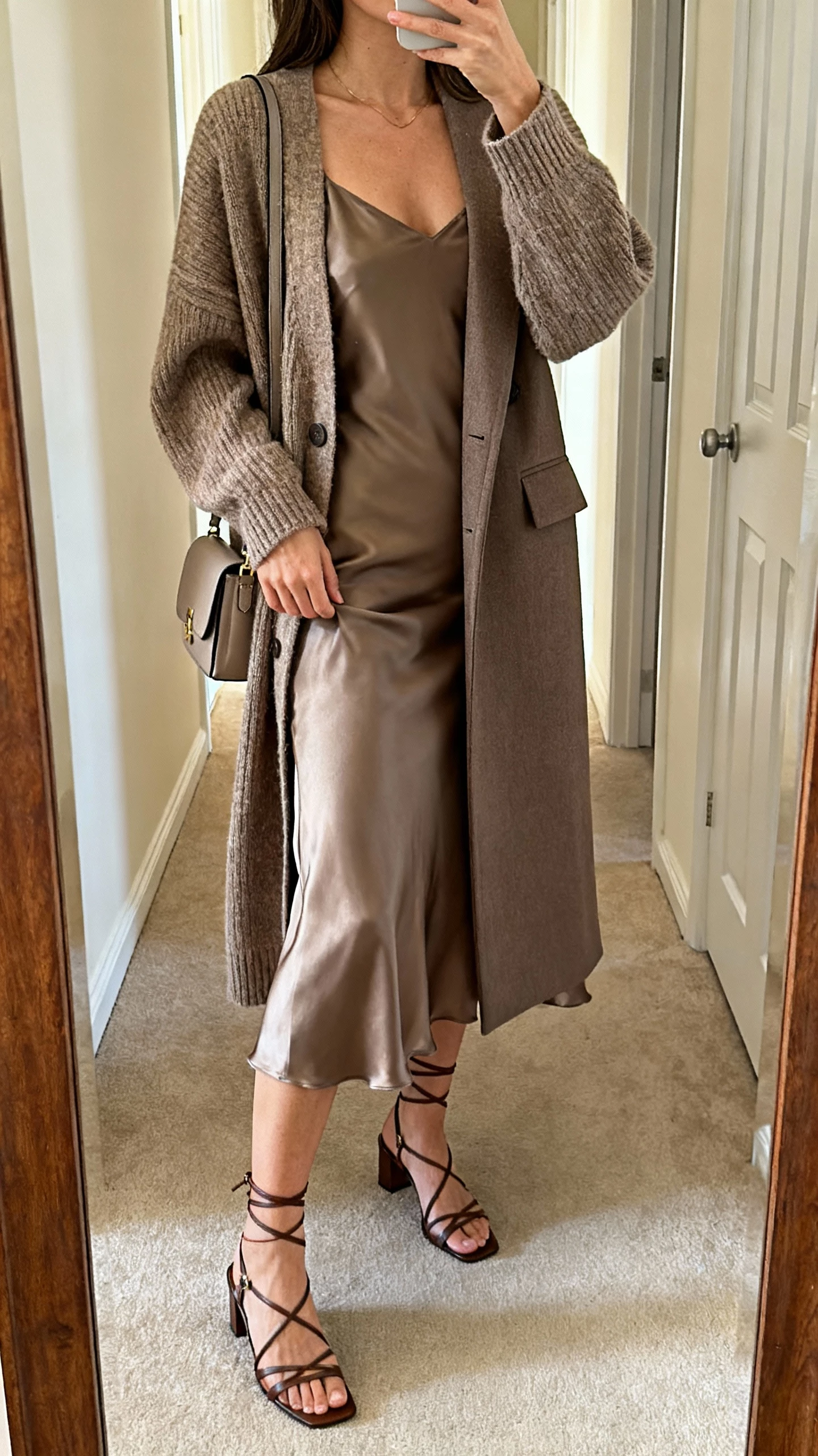 iPhone hallway mirror selfie of a woman in a luxe slip dress layered with an oversized cardigan and a tailored coat, strappy heels and a small shoulder bag, face not visible, cropped at neck, warm indoor lighting, natural iPhone photo quality.