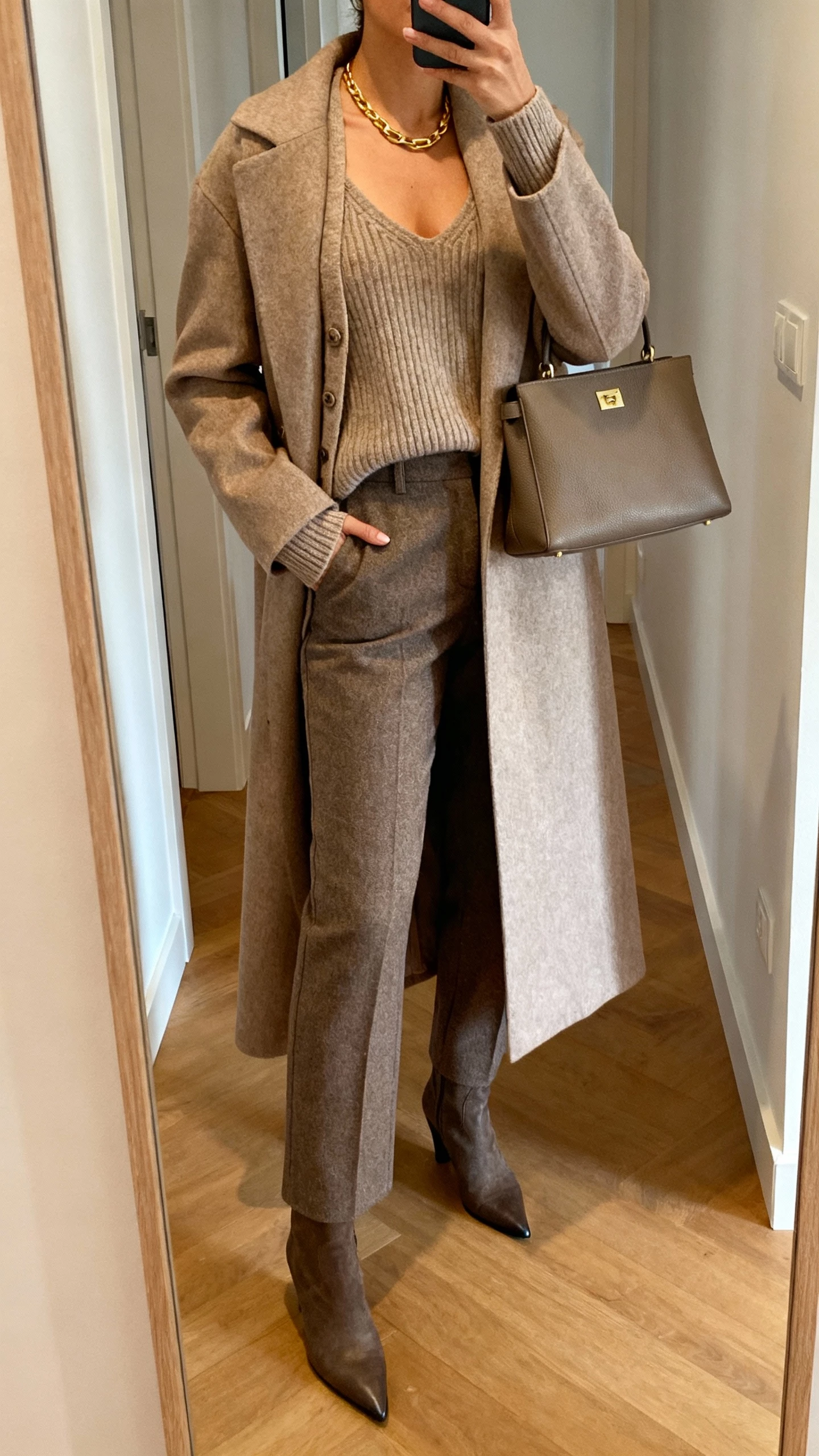 iPhone hallway mirror selfie of a woman in a matching knit sweater set (cardigan and tank) layered under a long maxi coat, tailored wool trousers, pointed-toe boots, simple gold necklace, structured handbag, face not visible, warm indoor lighting, natural iPhone photo quality.