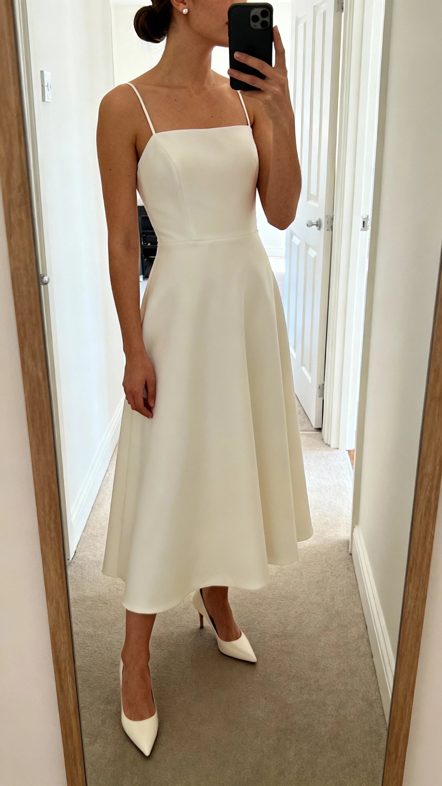 iPhone hallway mirror selfie of a woman in a modern minimalist A-line wedding gown with clean lines, smooth crepe, thin straps, no embellishments, sleek low bun, simple stud earrings, and pointed heels; face not visible, cropped at shoulders, bright indoor light, natural iPhone aesthetic.
