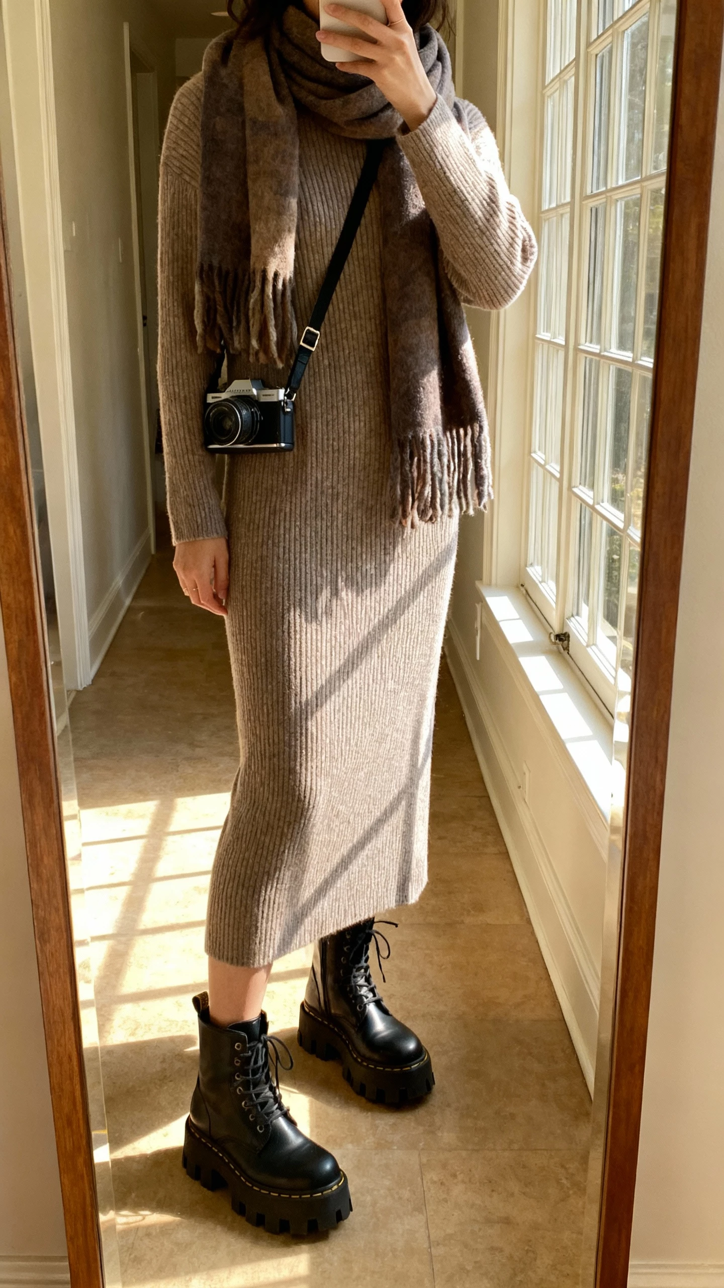 iPhone hallway mirror selfie of a woman in a ribbed sweater dress with chunky lug-sole boots, oversized scarf, crossbody camera bag, face not visible, cropped at shoulders, warm afternoon window light, natural iPhone photo quality.