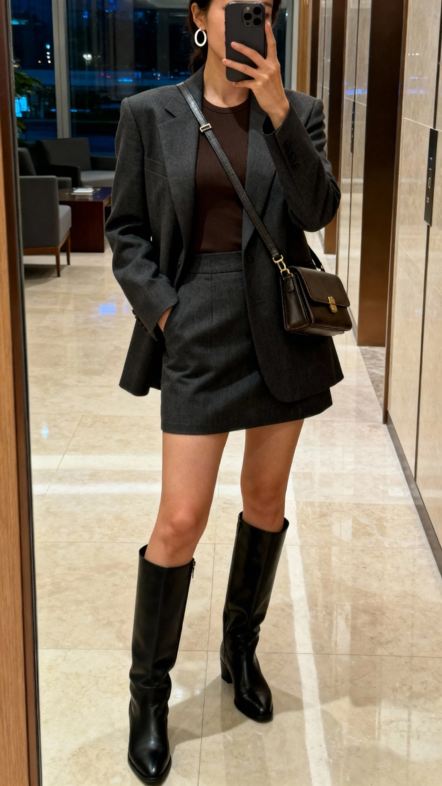 iPhone hallway mirror selfie of a woman in a tailored blazer, mini skirt, fitted top, knee-high boots, simple hoop earrings, and a structured crossbody, face not visible, office lobby setting, indoor evening lighting, natural iPhone photo quality