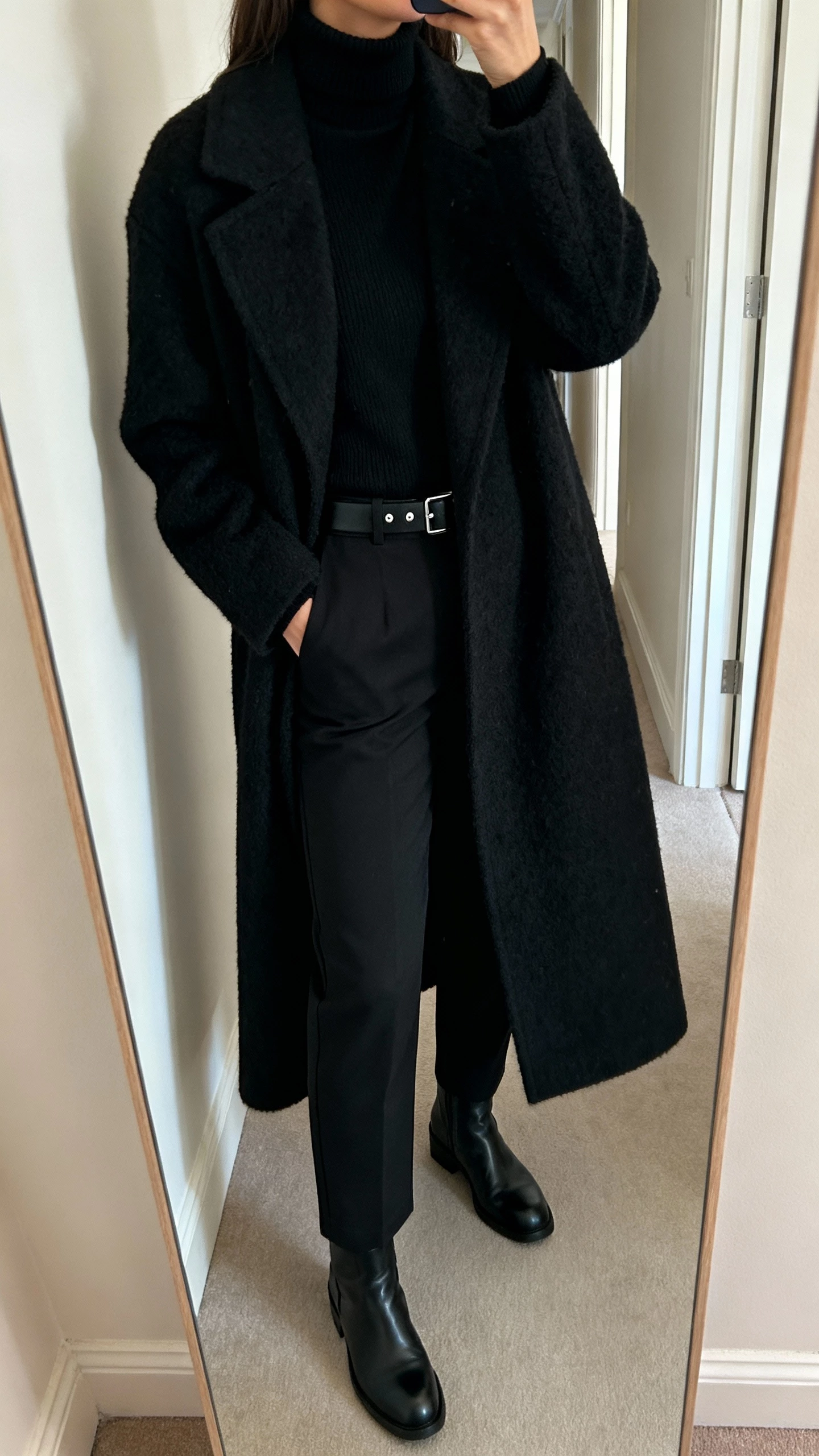 iPhone hallway mirror selfie of a woman in an all-black layered look: black turtleneck, black long wool coat, black straight trousers, black leather boots, slim black belt, minimal silver studs, face not visible, cropped at neck, natural indoor light, iPhone photo.
