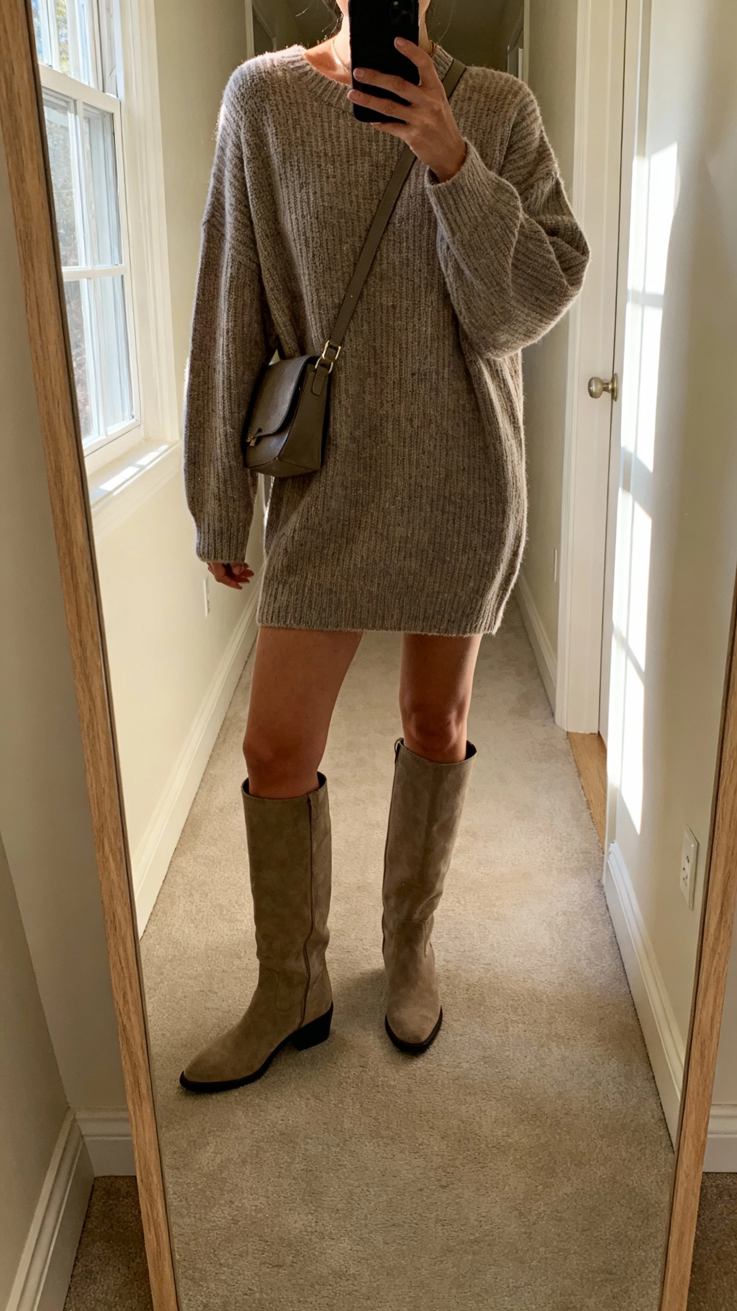 iPhone hallway mirror selfie of a woman in an oversized knit dress with knee-high boots and a crossbody bag, face not visible, cropped at shoulders, soft afternoon window light, natural iPhone photo quality.