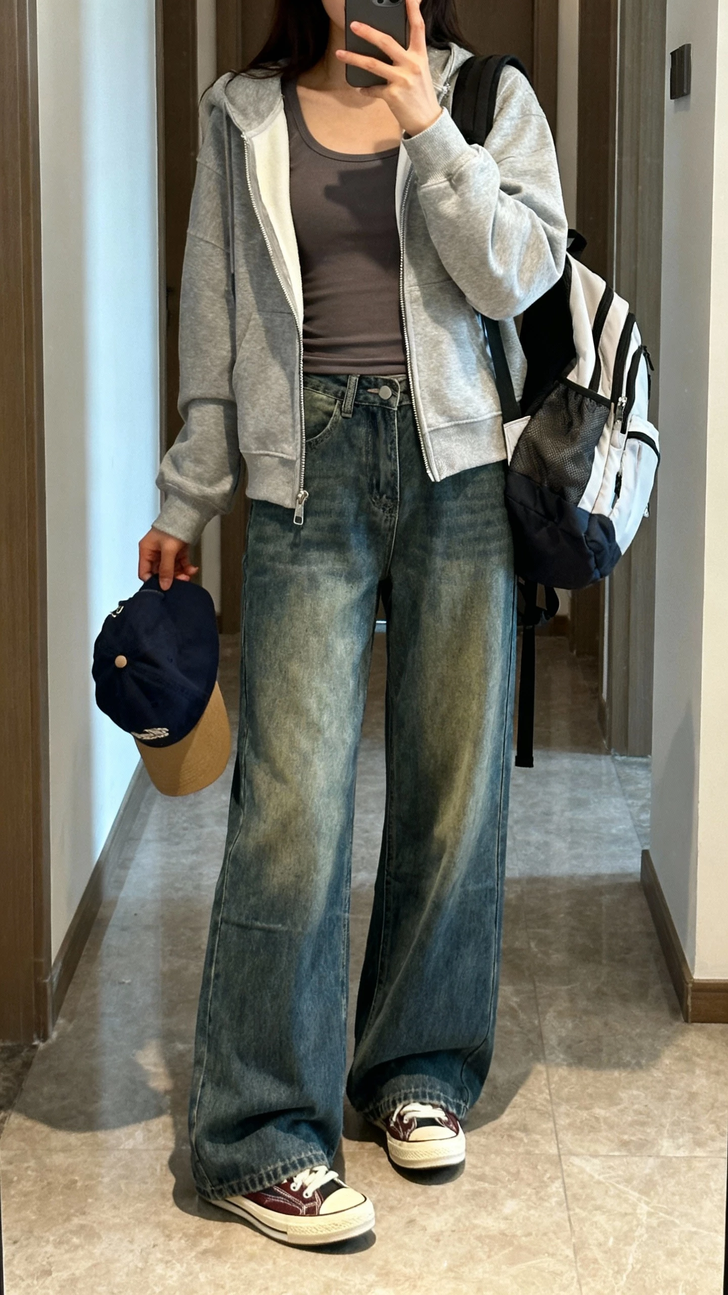 iPhone hallway mirror selfie of a woman in wide leg jeans, zip hoodie slightly open over a fitted tee, retro sneakers, baseball cap in hand, sporty backpack, face not visible, daytime natural light, natural iPhone photo quality.