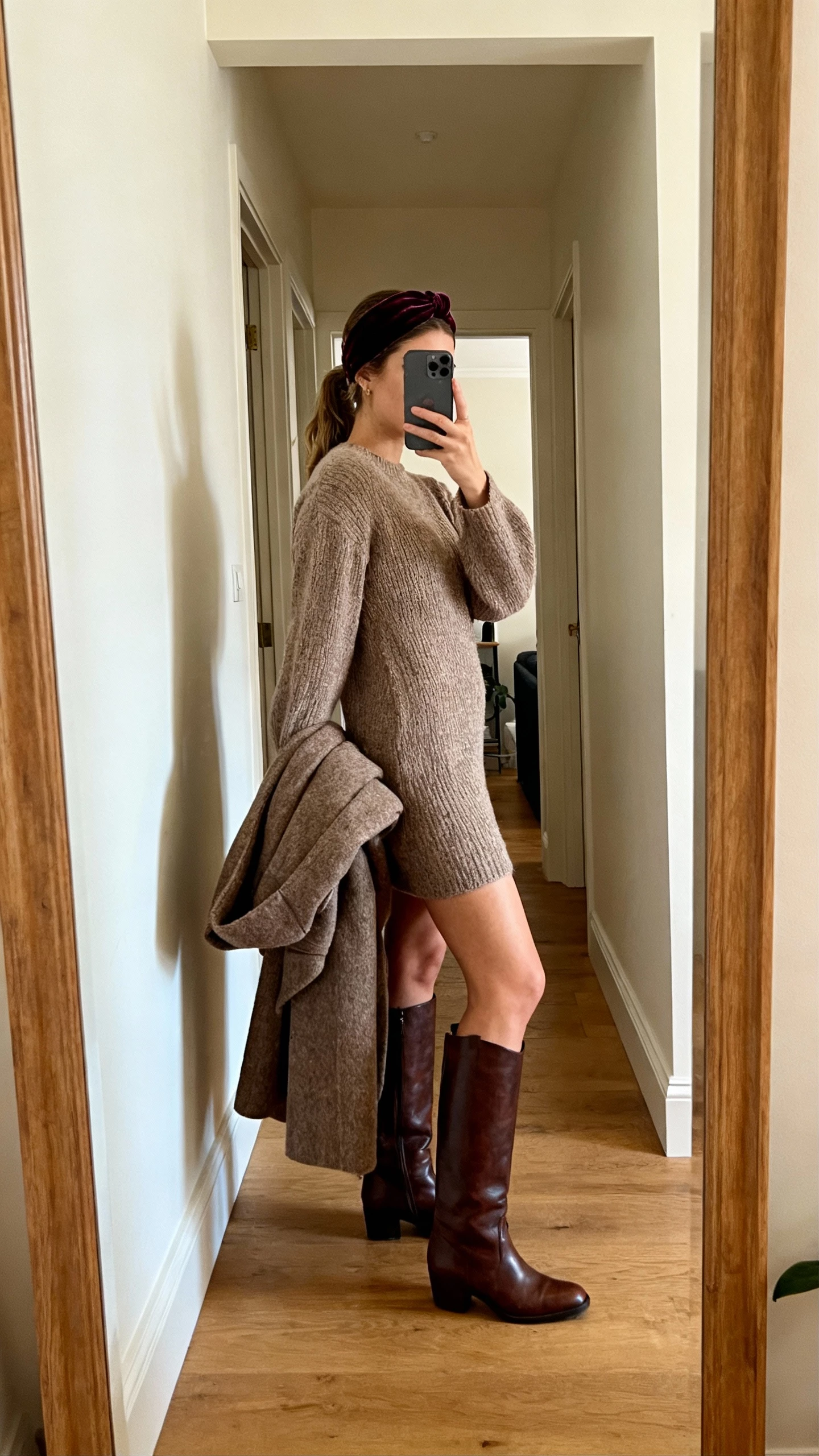 iPhone hallway mirror selfie of a woman wearing a velvet headband, cozy sweater dress, tall leather boots, and a wool coat draped over arm, face not visible, side angle, warm indoor lighting, natural iPhone photo quality.