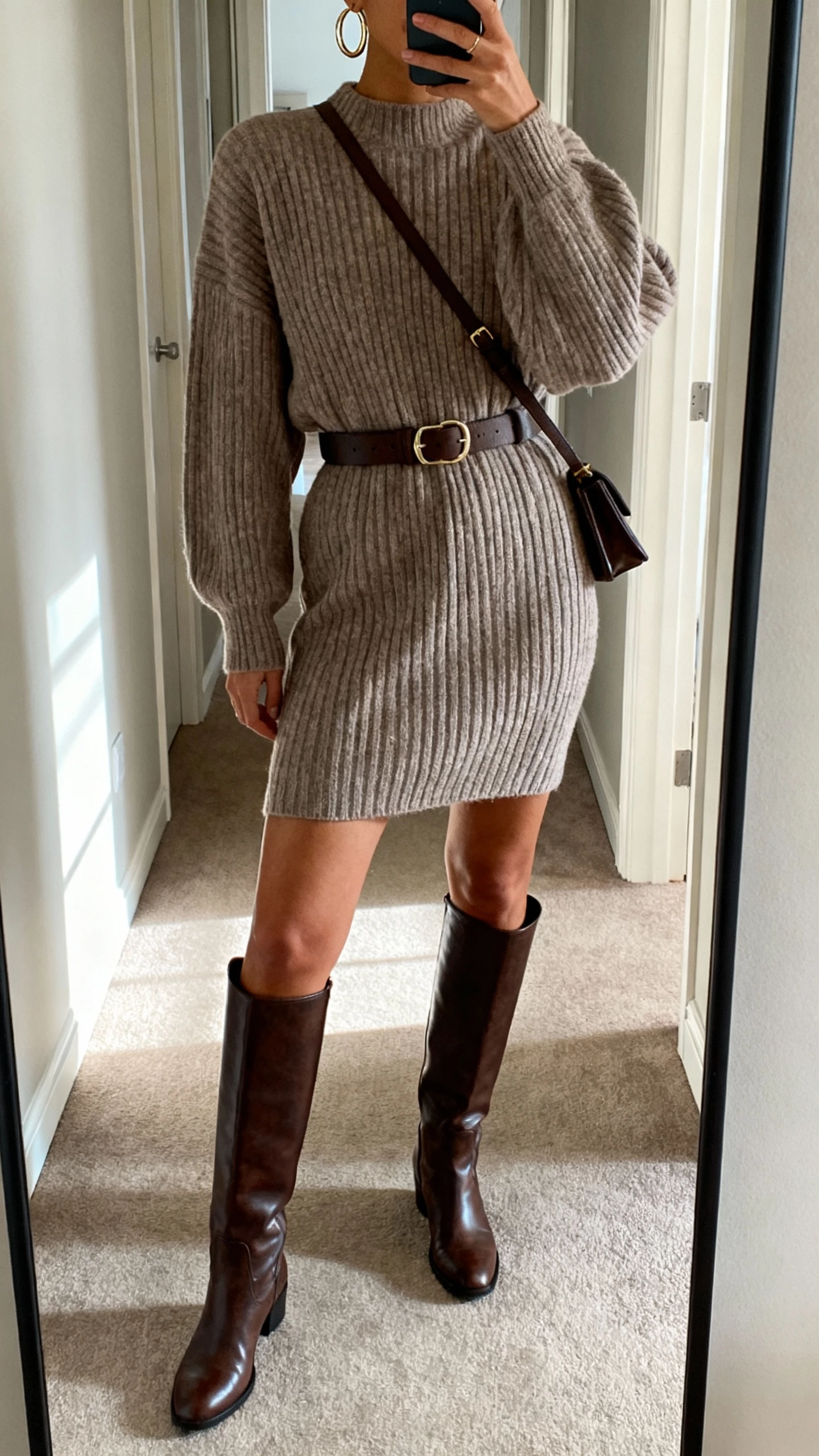 iPhone hallway mirror selfie of a woman wearing a ribbed knit sweater dress with tall knee-high leather boots, belt at the waist, hoop earrings, crossbody bag, face not visible, cropped at shoulders, natural morning light, realistic iPhone photo