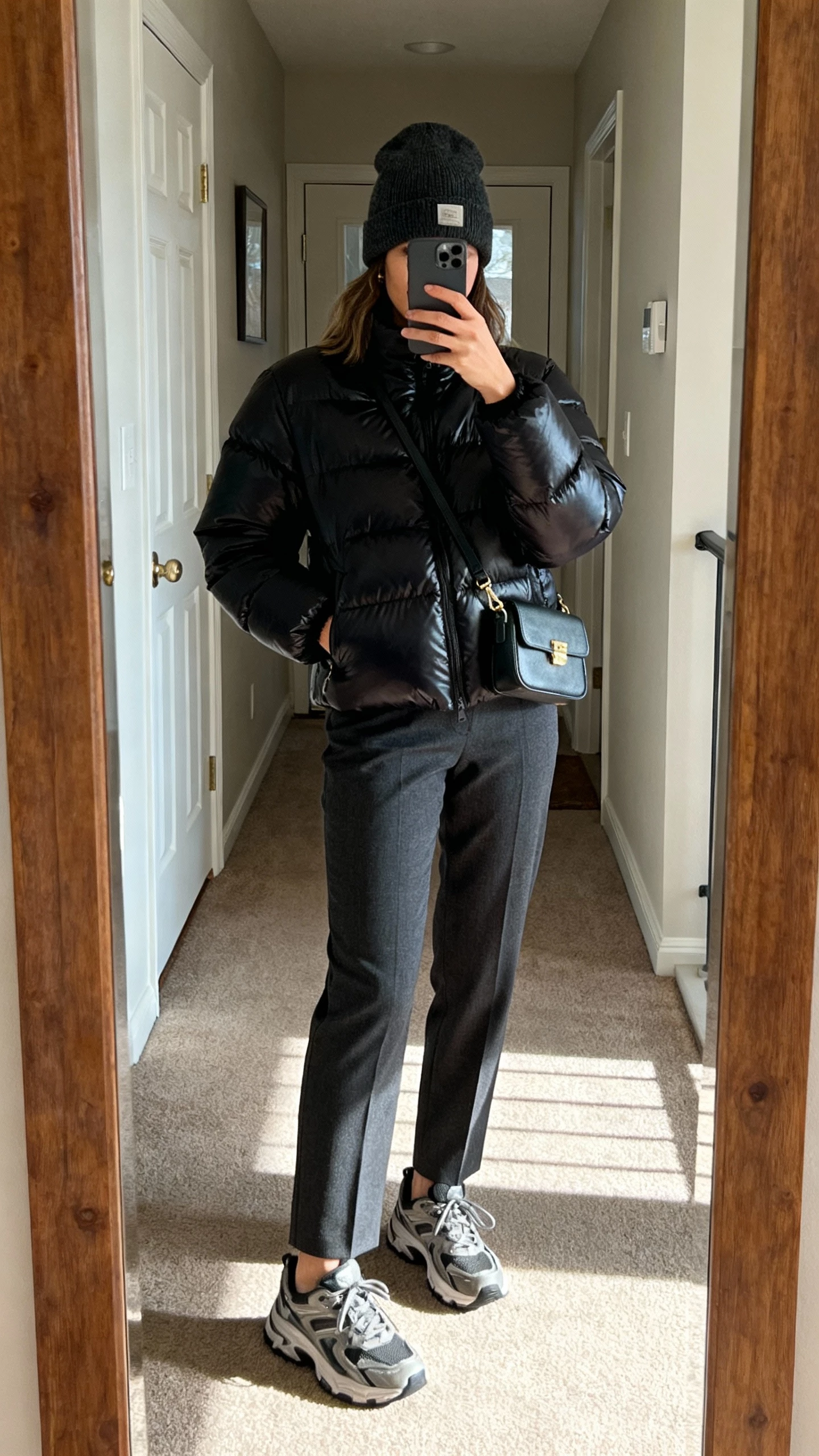 iPhone hallway mirror selfie of a woman wearing a matte black puffer jacket with tailored charcoal trousers, chunky sneakers, beanie, mini shoulder bag, face not visible, full-length mirror in apartment entryway, afternoon natural light, casual iPhone photo