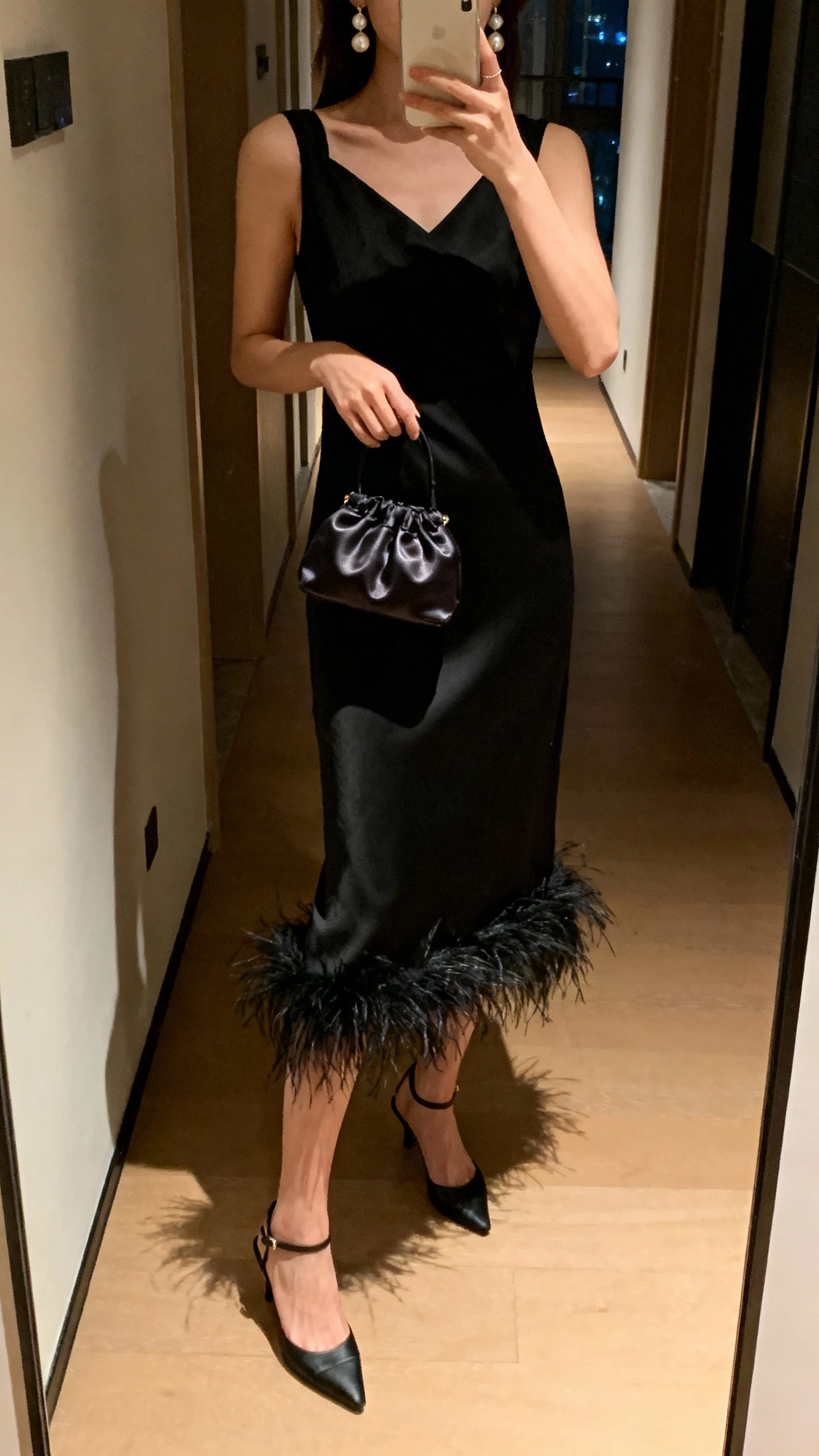 iPhone hallway mirror selfie of a woman wearing a black feather-trim midi dress with slingback heels, pearl drop earrings, satin mini bag, face not visible, cropped at shoulders, evening indoor lighting, natural iPhone photo quality.