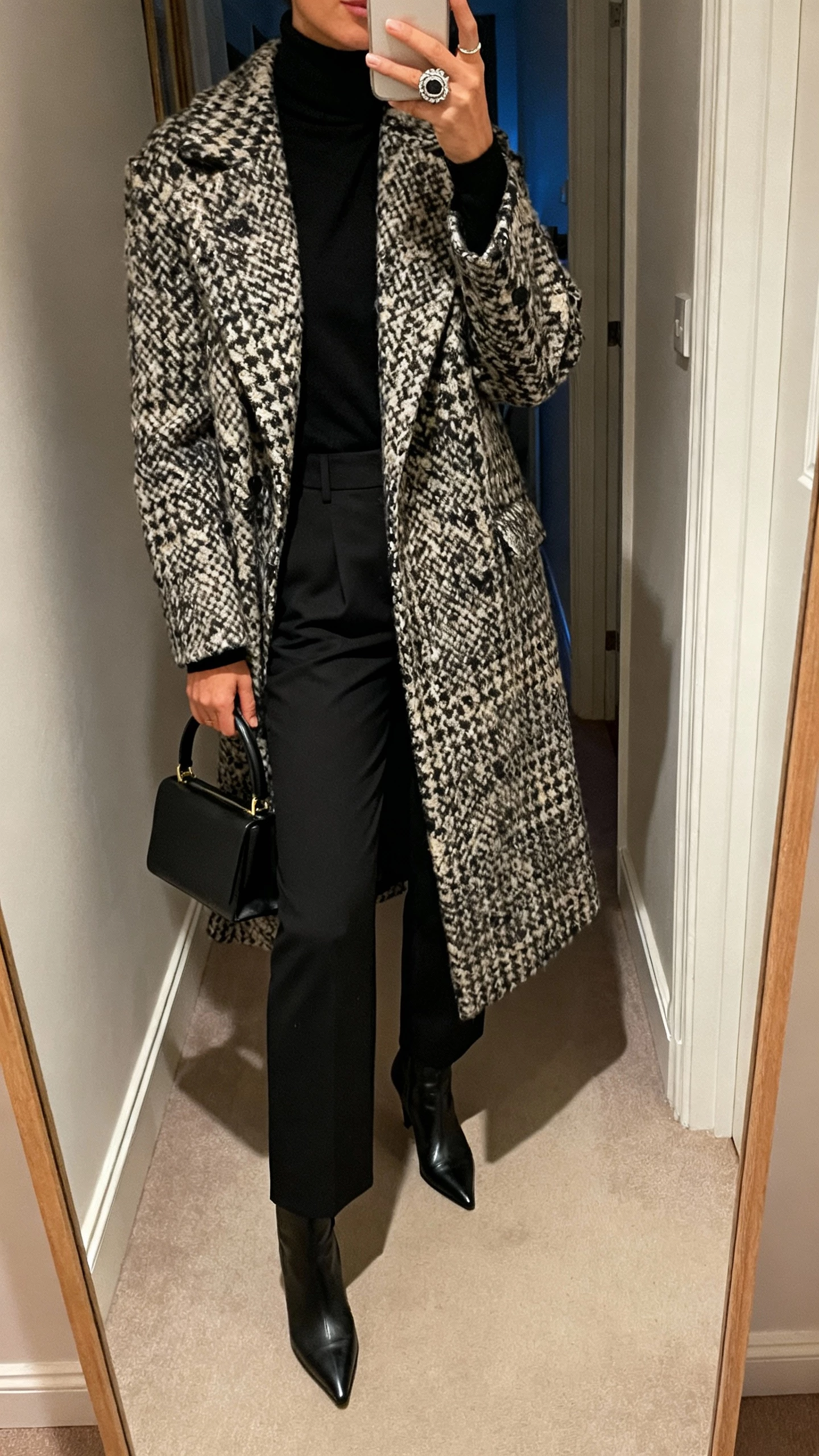 iPhone hallway mirror selfie of a woman wearing a bold statement coat over a monochrome black base (turtleneck and tailored pants), pointed ankle boots, top-handle bag, chunky ring, face not visible, cropped at neck, soft evening indoor lighting, natural iPhone aesthetic.