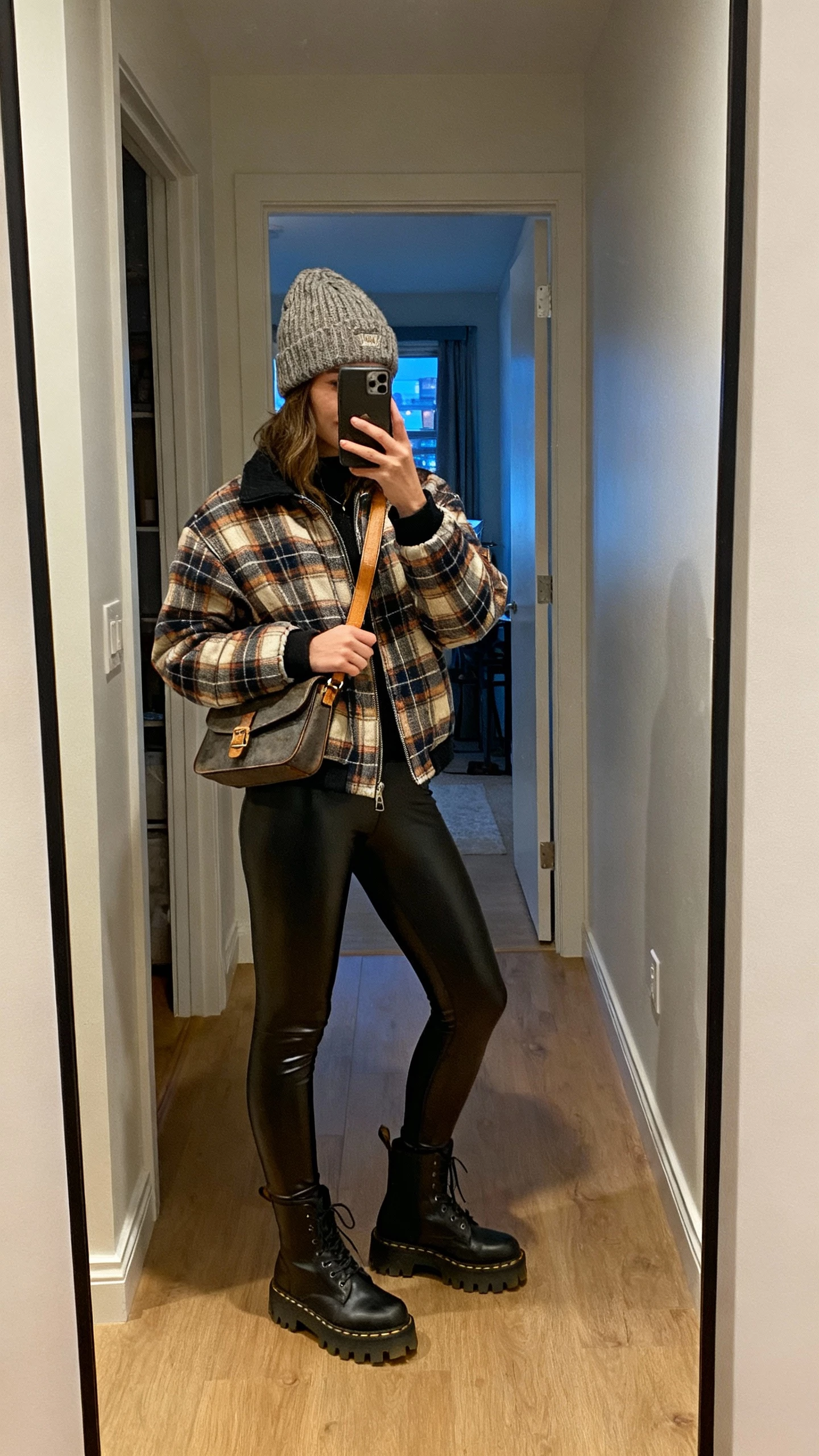 iPhone hallway mirror selfie of a woman wearing a plaid shacket with faux-leather leggings, lug-sole boots, crossbody, knit beanie, face not visible, apartment hallway mirror, evening indoor lighting, natural iPhone photo quality