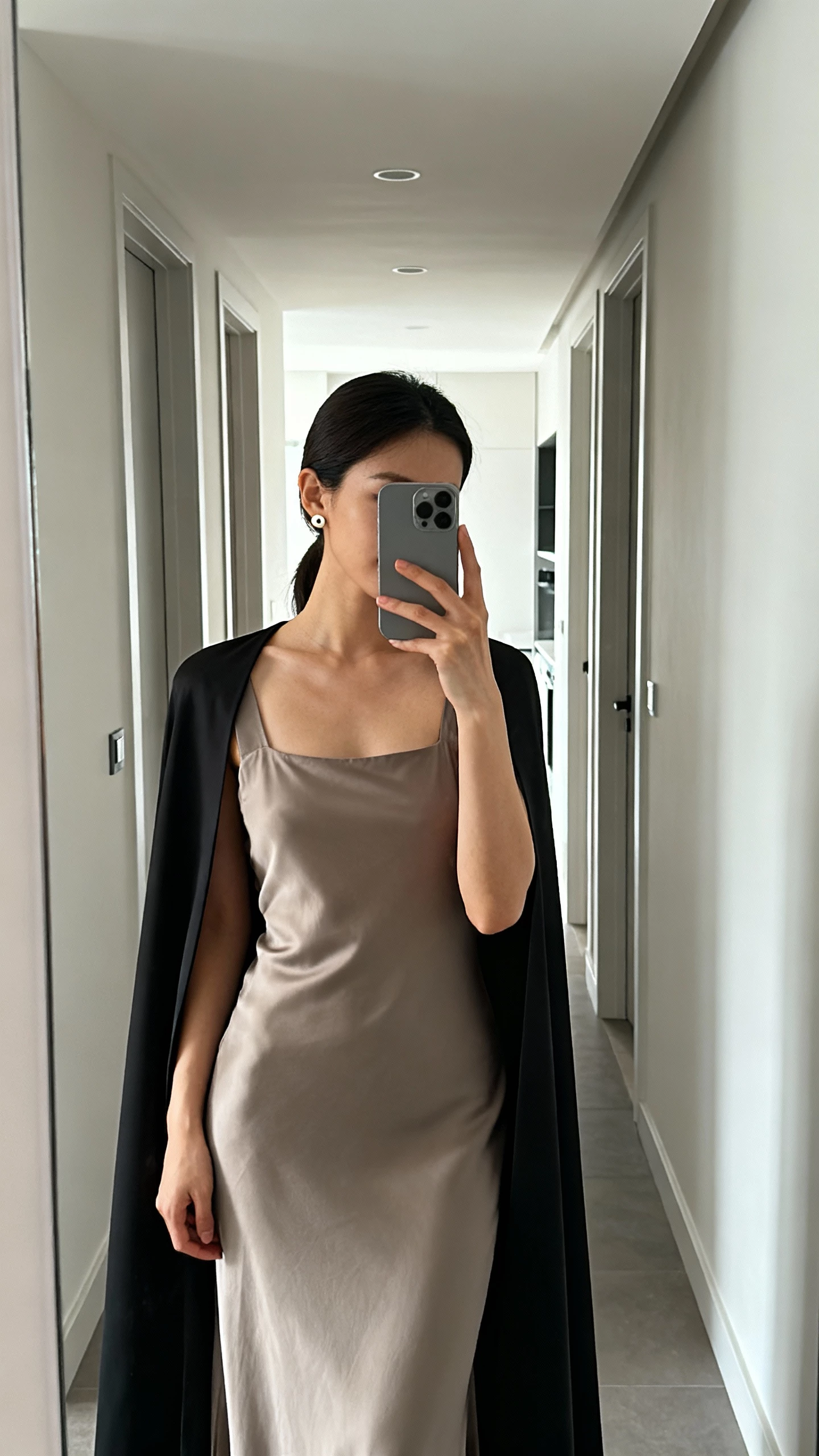iPhone hallway mirror selfie of a woman wearing a square-neck slip dress with a streamlined cape, simple stud earrings, face not visible, modern apartment corridor, diffused daylight, natural iPhone photo