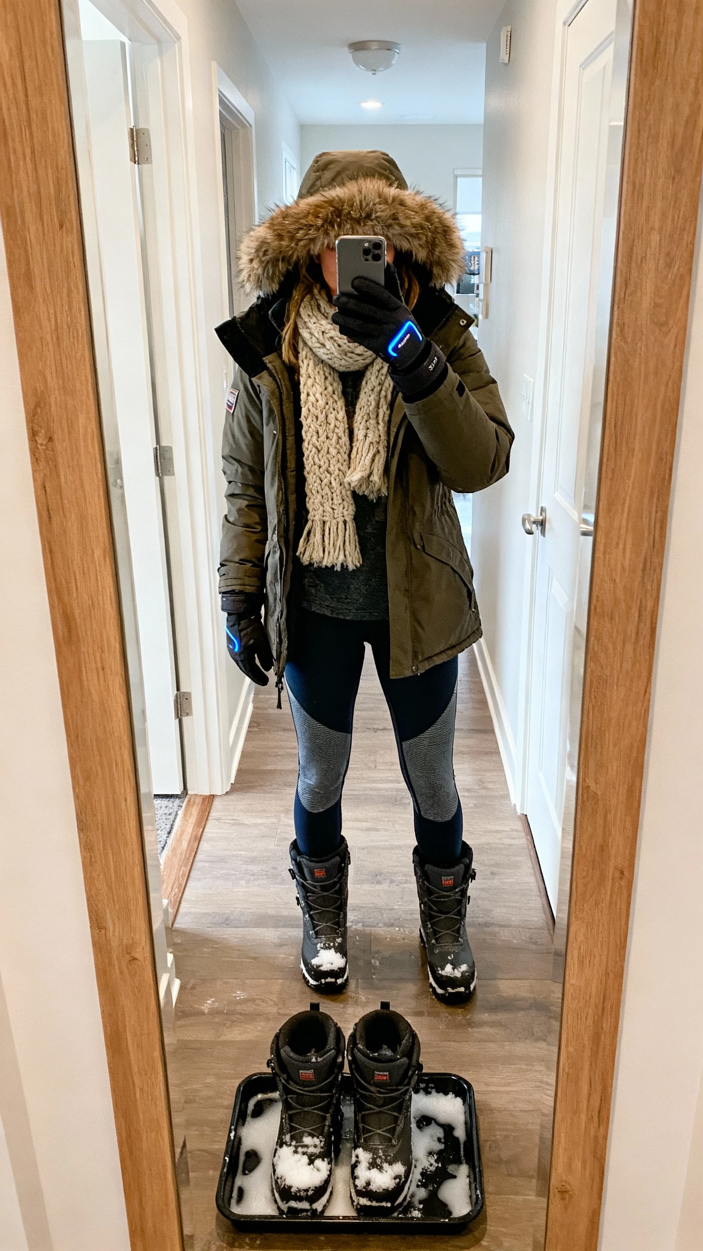 iPhone hallway mirror selfie of a woman wearing waterproof snow boots, insulated parka with faux-fur hood, thermal joggers, chunky scarf, and tech gloves, face not visible, apartment entryway with melting snow boots tray, natural daylight, iPhone photo quality.