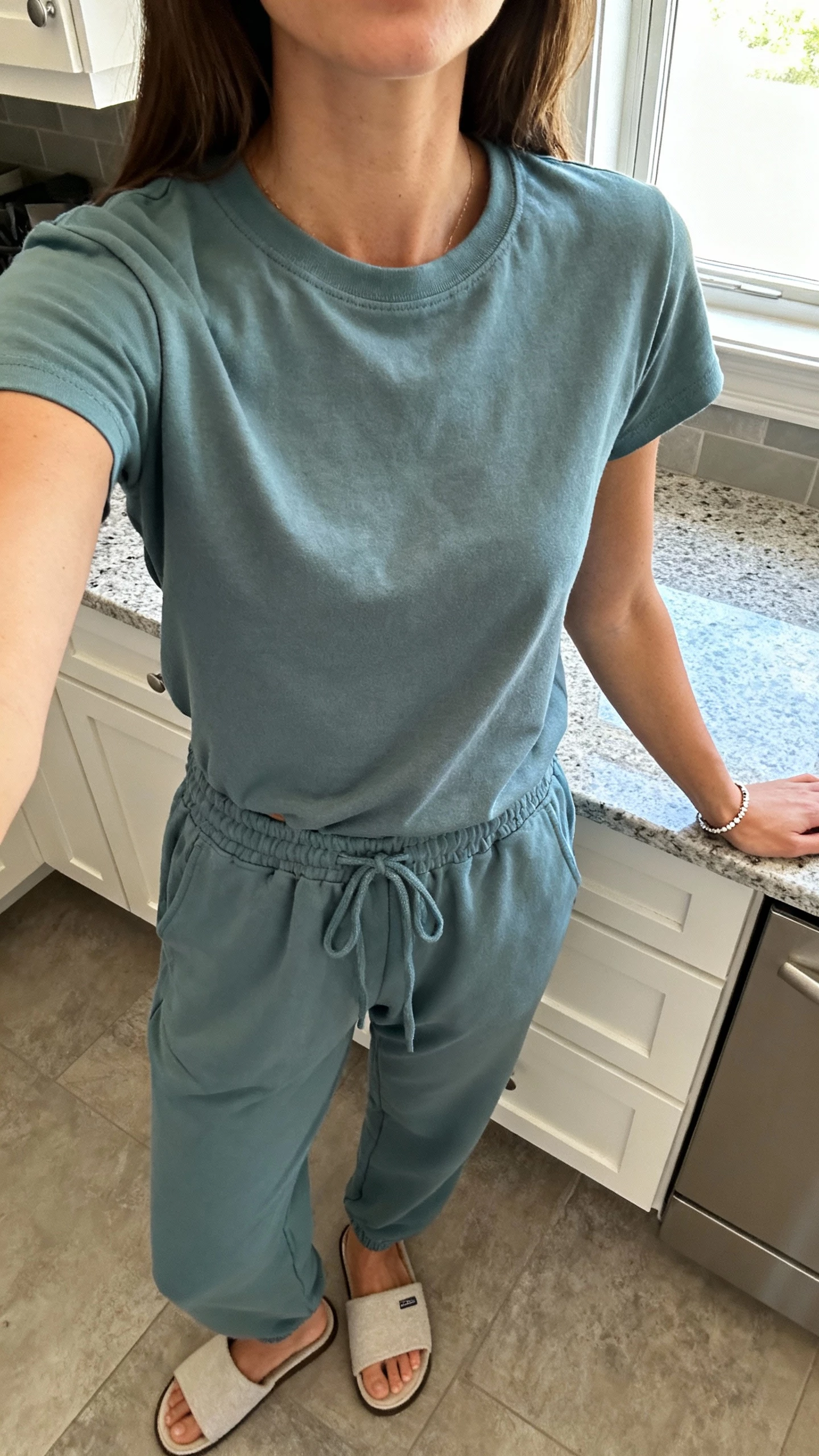 iPhone kitchen counter selfie cropped at neck showing a woman in a jersey tee and drawstring pants set with minimal bracelet and house slides, face not visible, bright kitchen window light, natural iPhone photo quality.