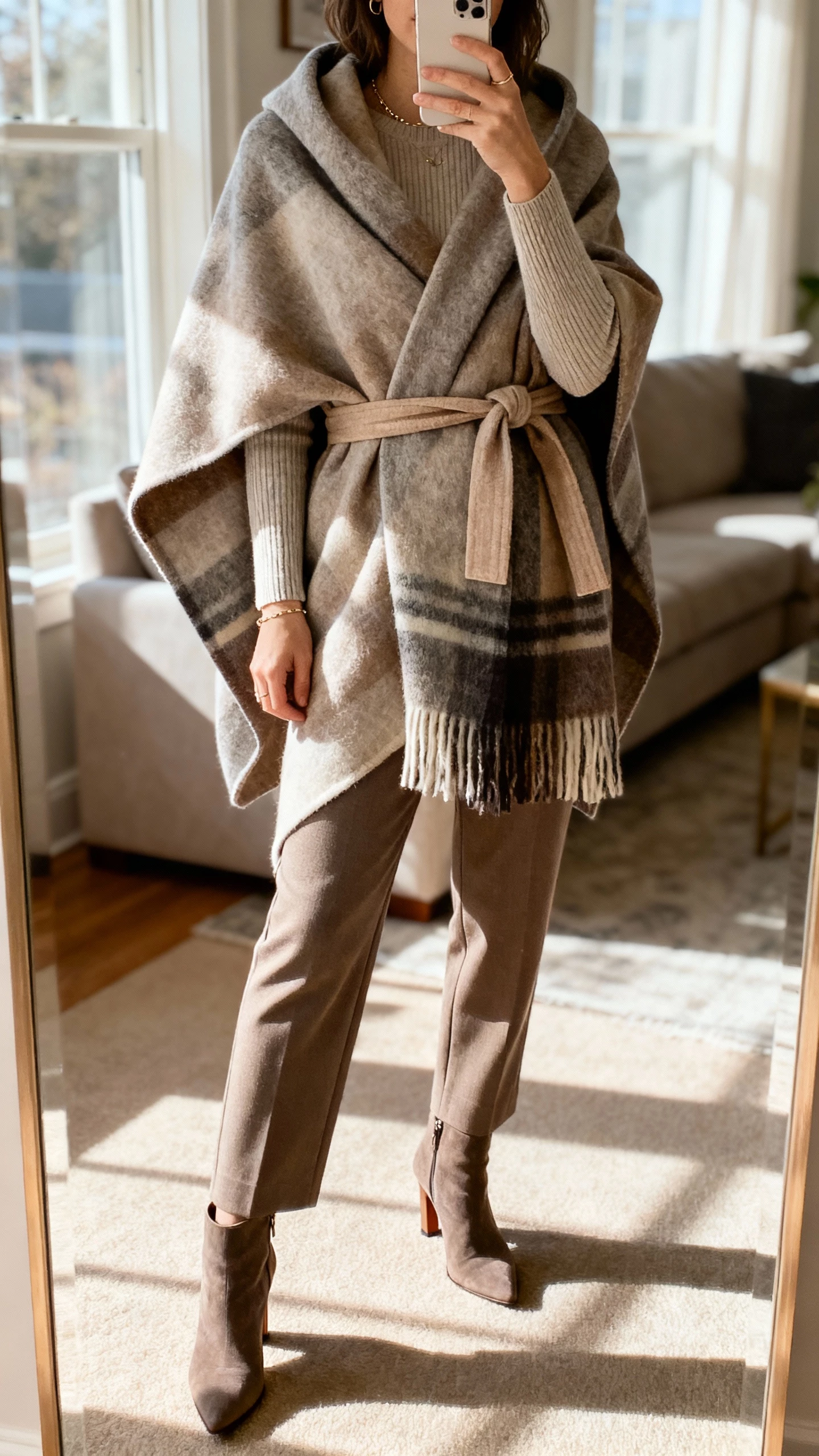 iPhone living room mirror selfie of a woman in a belted wrap cape styled from a blanket scarf, long-sleeve knit, straight-leg trousers, heeled booties, minimal jewelry, face not visible, afternoon window light, natural iPhone photo.