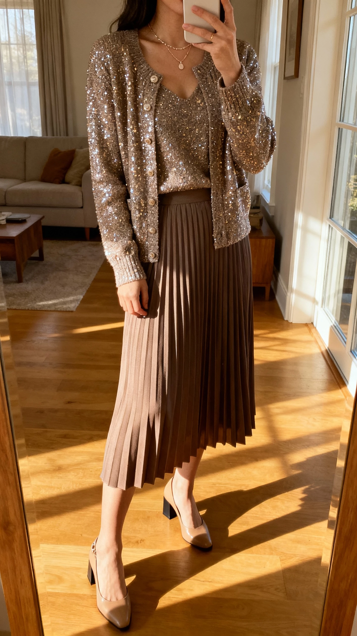 iPhone living room mirror selfie of a woman in a pleated midi skirt with a sparkly cardigan set, dainty necklace, and block heels, face not visible, warm afternoon window light, natural iPhone photo quality.