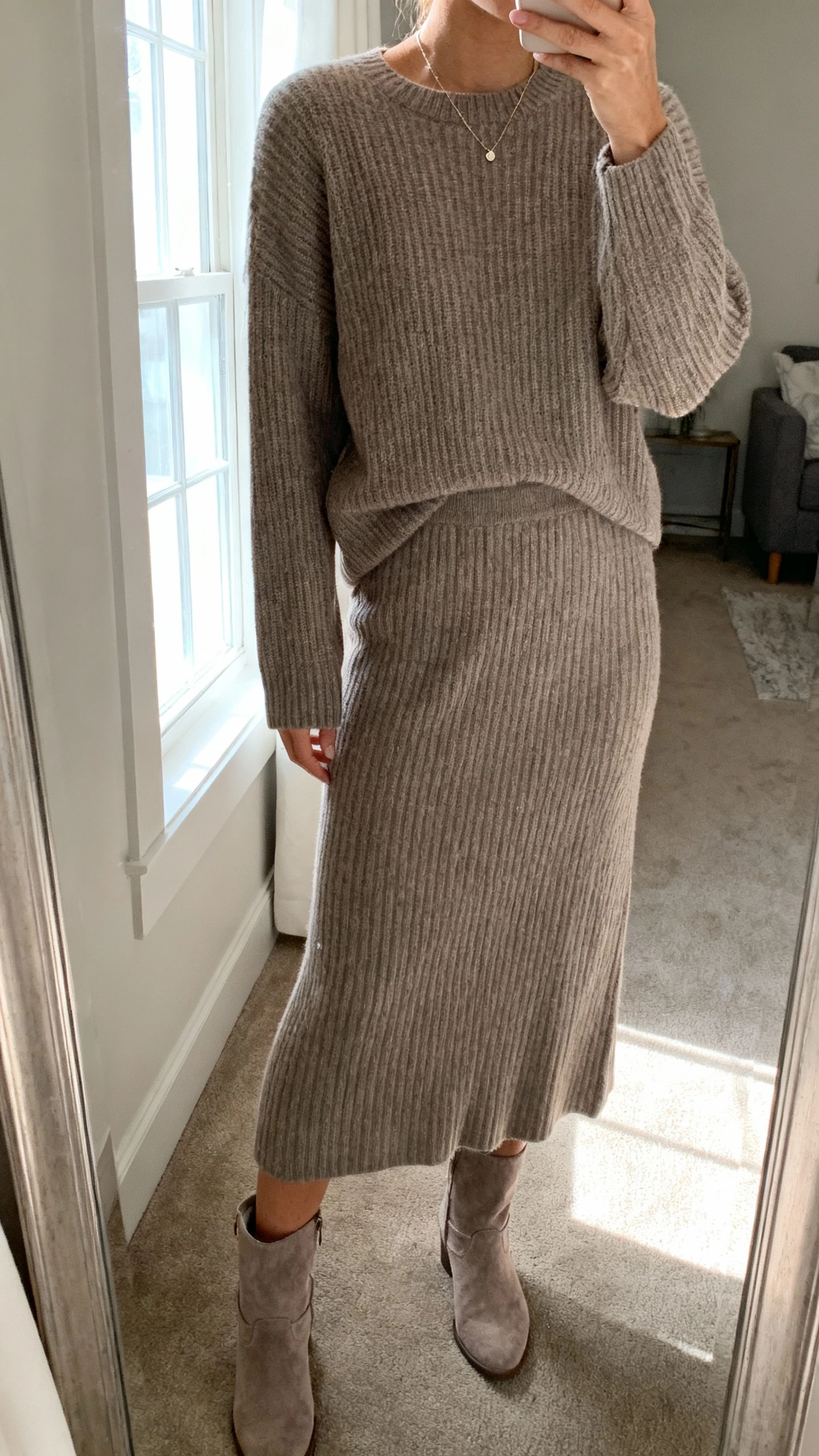iPhone living-room mirror selfie of a woman wearing a monochrome knit set: taupe ribbed sweater with matching knit midi skirt, tonal suede boots, delicate necklace, face not visible, cropped at neck, window light, natural iPhone photo quality.