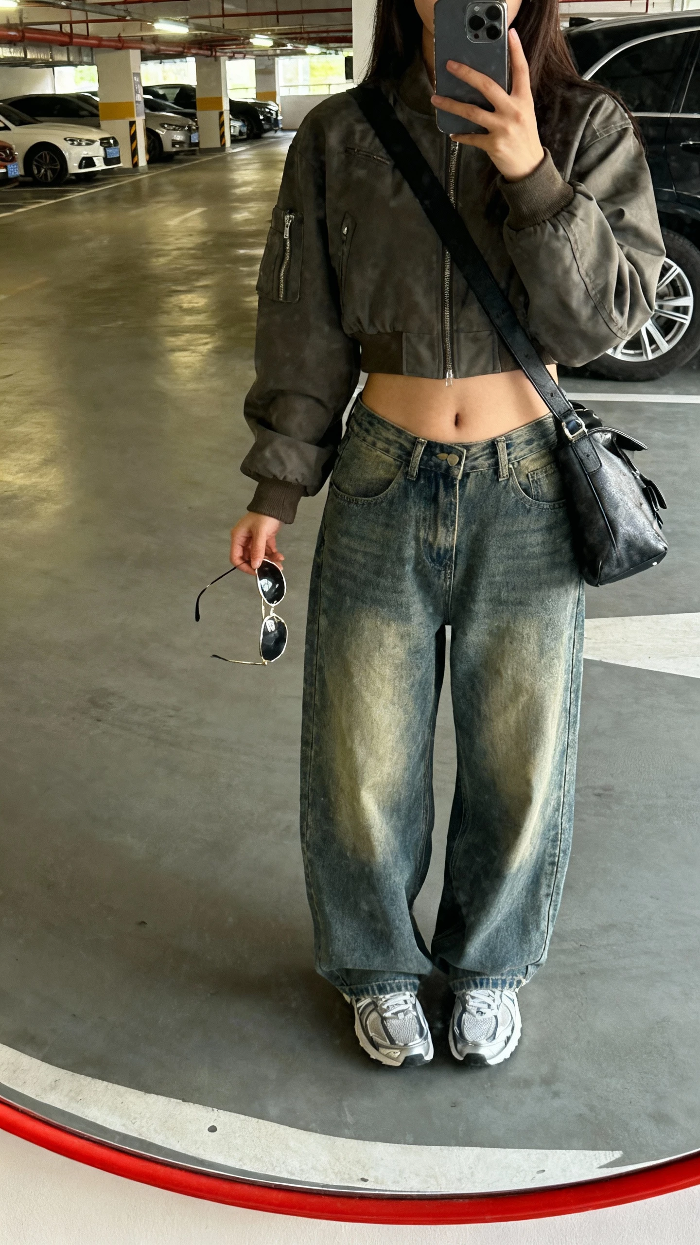 iPhone low-angle mirror selfie of a woman in a cropped bomber jacket, wide baggy jeans, sleek sneakers, crossbody bag, aviator-style sunglasses in hand; parking garage mirror, face not visible, natural daylight, casual iPhone photo.