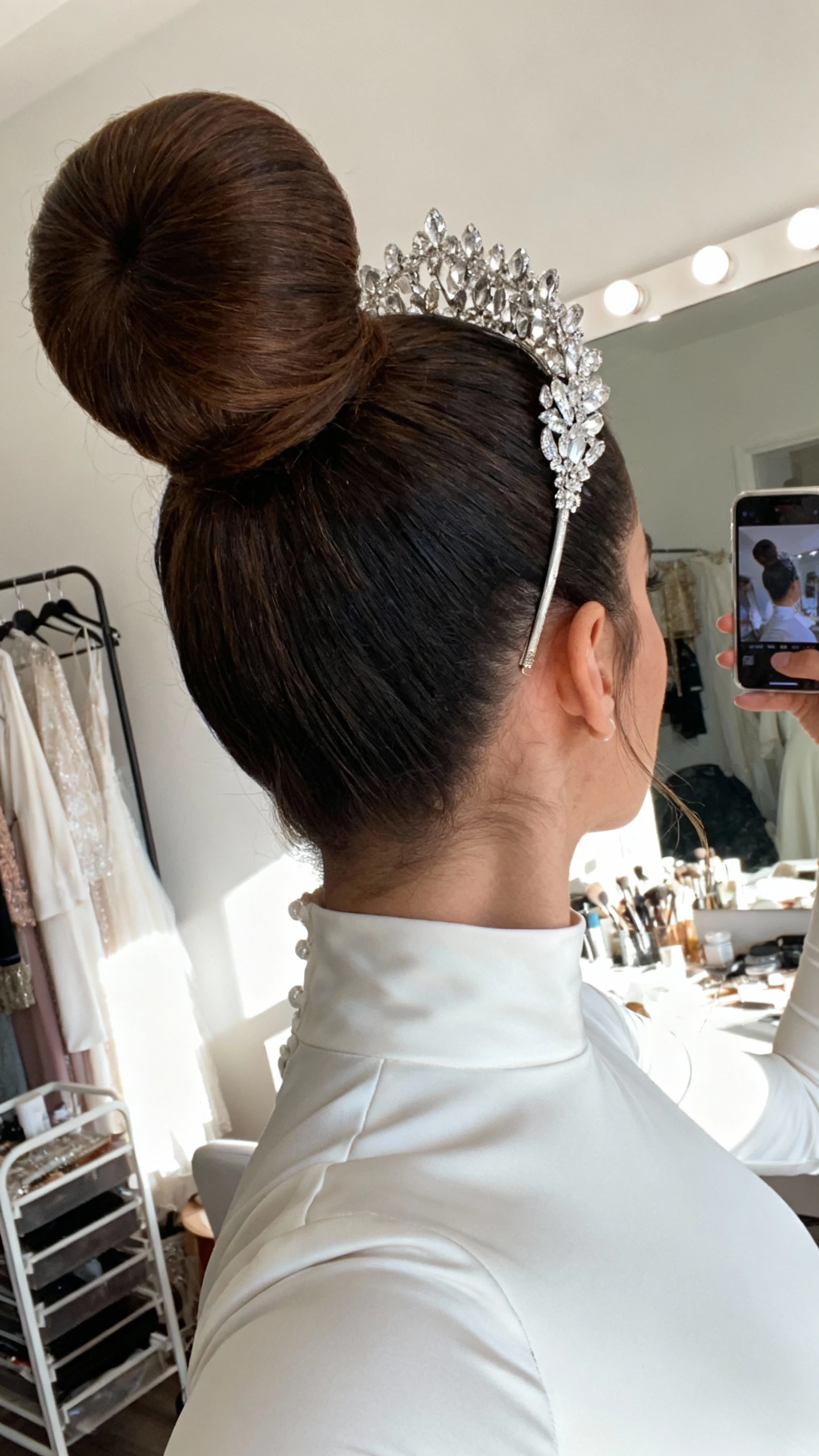 iPhone low-angle mirror selfie of a woman with a high ballet bun with dramatic height, statement crystal headband, sleek high‑neck bridal dress; face not visible, studio dressing area, bright natural daylight, natural iPhone photo quality.