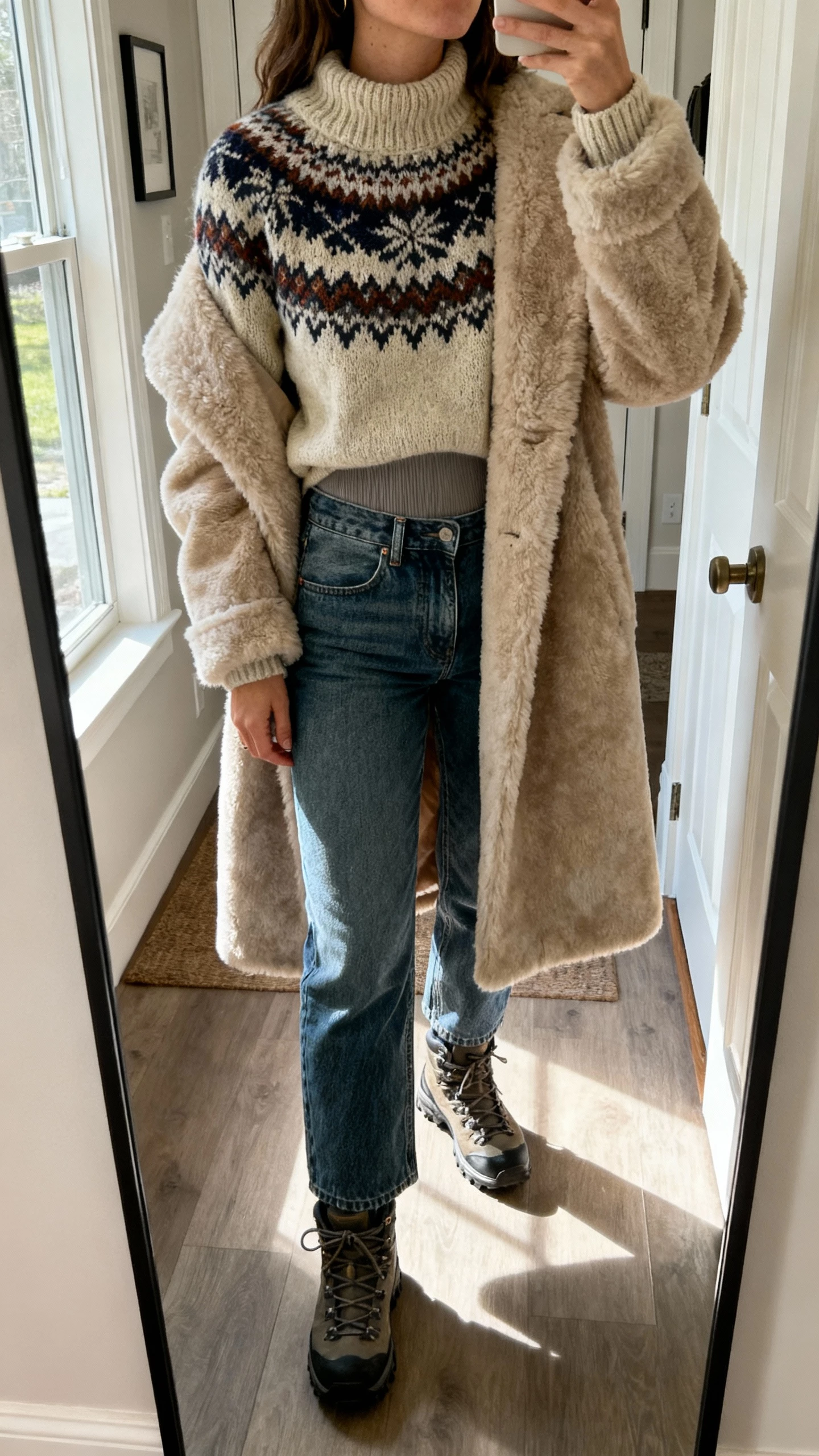 iPhone mirror selfie cropped at neck of a woman in a chunky Fair Isle knit over a thermal crew base layer, high-rise straight-leg jeans, a mid-length shearling coat, and rugged hiker boots; face not visible, apartment entryway mirror, afternoon window light, natural iPhone photo quality.