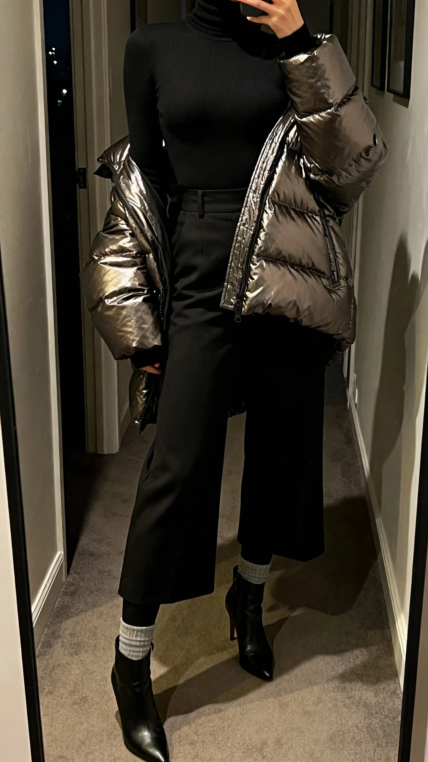 iPhone mirror selfie cropped at neck of a woman in a form-fitting black turtleneck, high-waist wide-leg trousers with thermal leggings underneath, sleek heeled ankle boots, and a bold statement coat (metallic puffer) draped over shoulders; dim hallway mirror before a night out, indoor lighting, face not visible, natural iPhone photo quality.