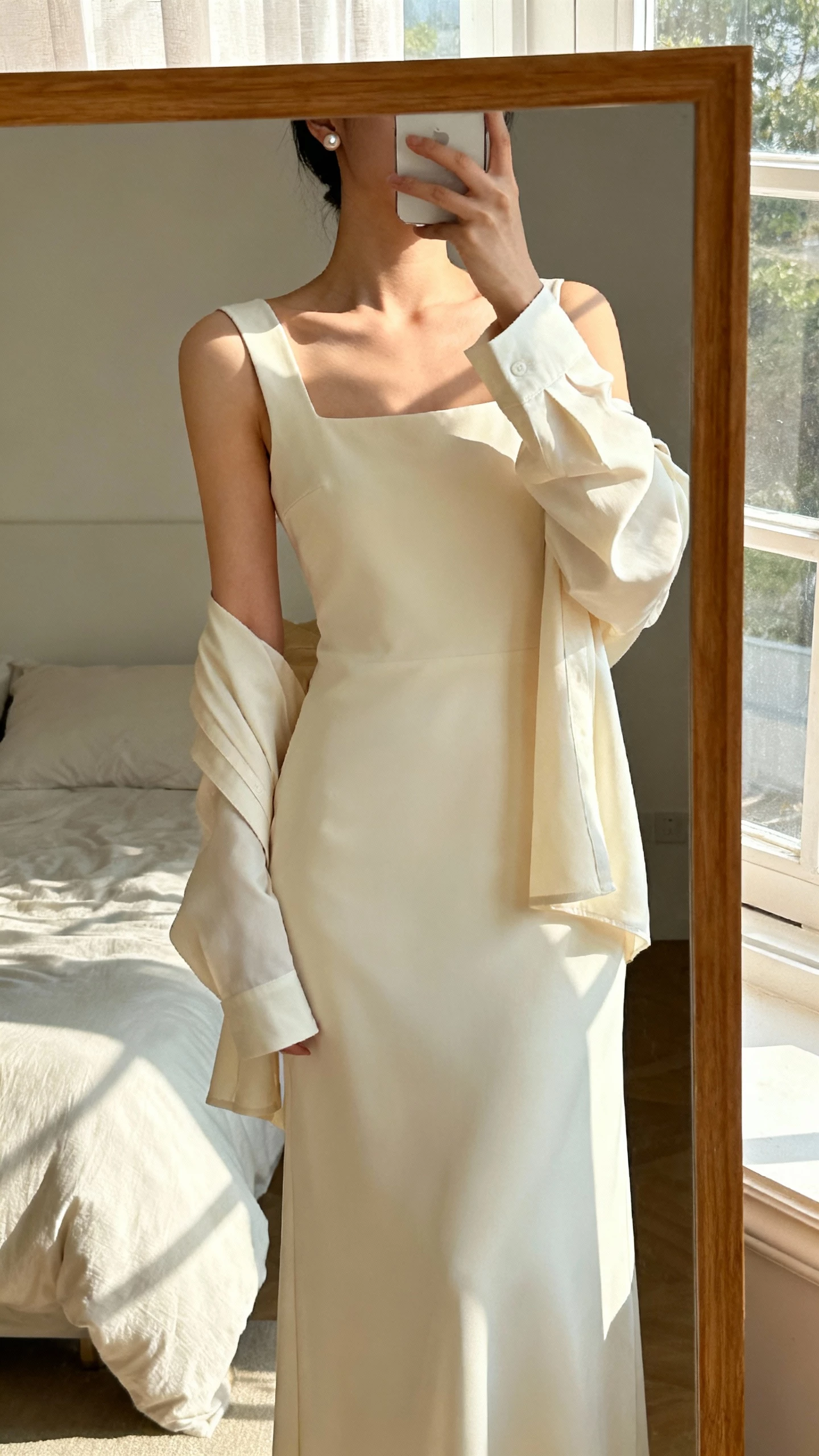 iPhone mirror selfie cropped at neck of a woman in a contemporary square-neck gown with detachable sleeves (shown on), sleek silhouette, simple studs, face not visible, minimalist bedroom mirror, soft morning window light, natural iPhone photo quality